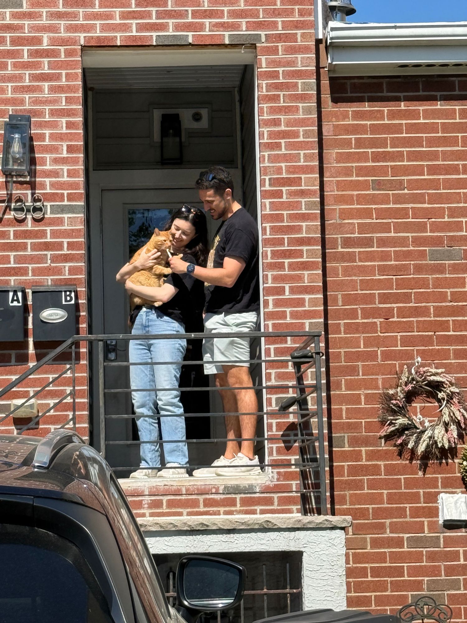 Couple holding an orange cat on a brick townhouse porch steps in sunny weather.