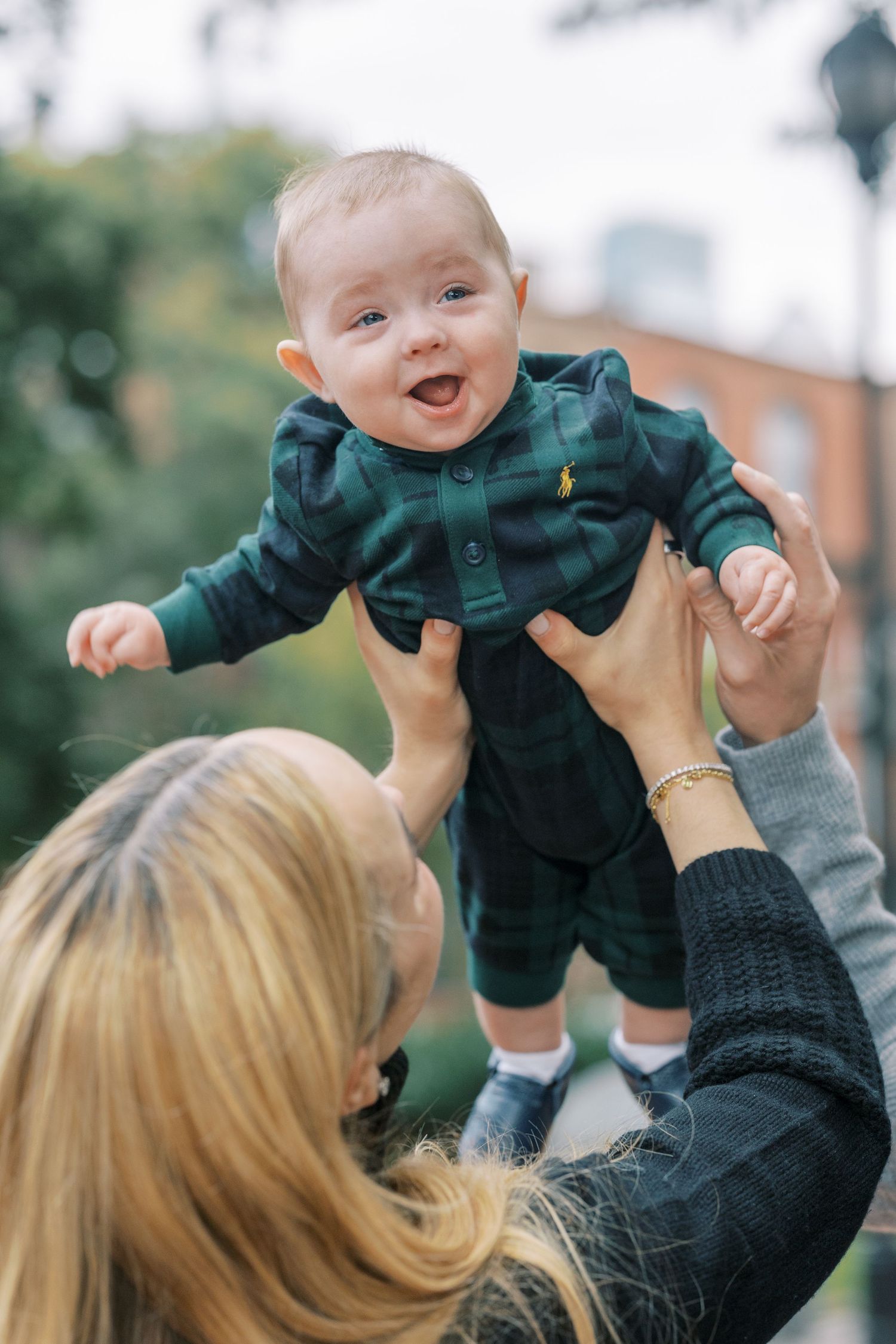 A joyful moment of a baby in a green outfit being lifted up into the air against a blurred outdoor background.