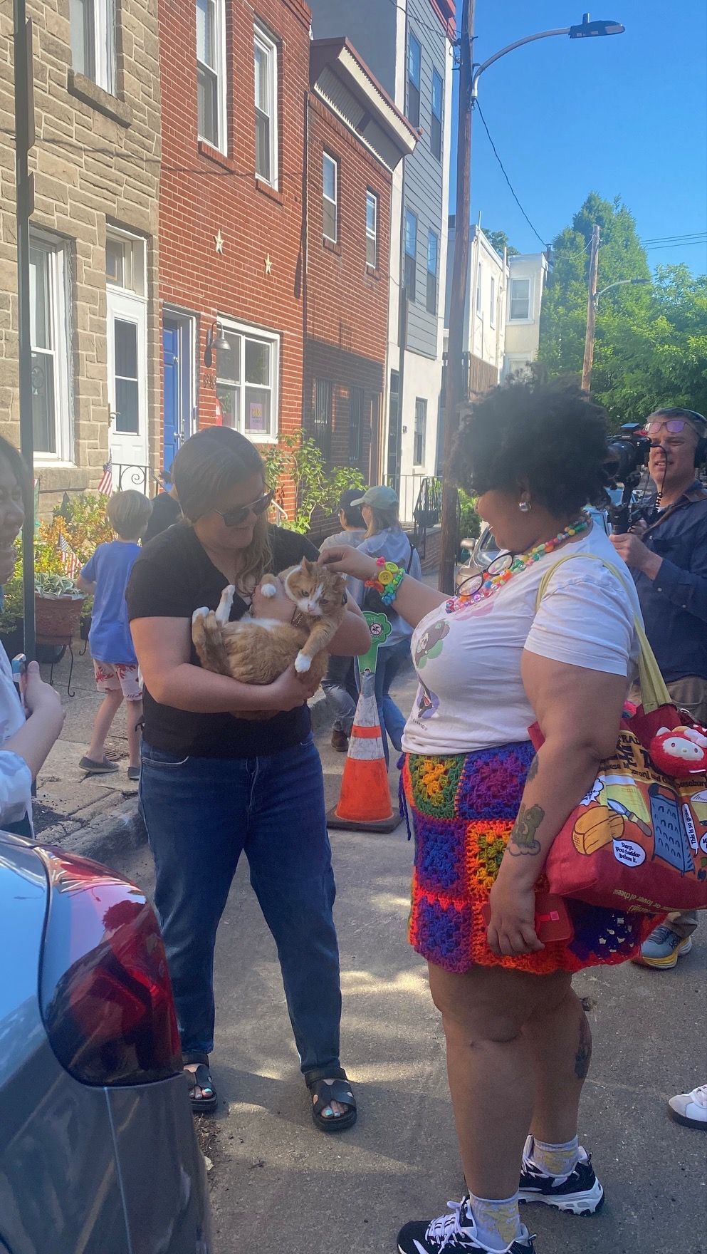 Person holding an orange cat while talking to a woman in colorful crochet skirt on a sunny urban street.