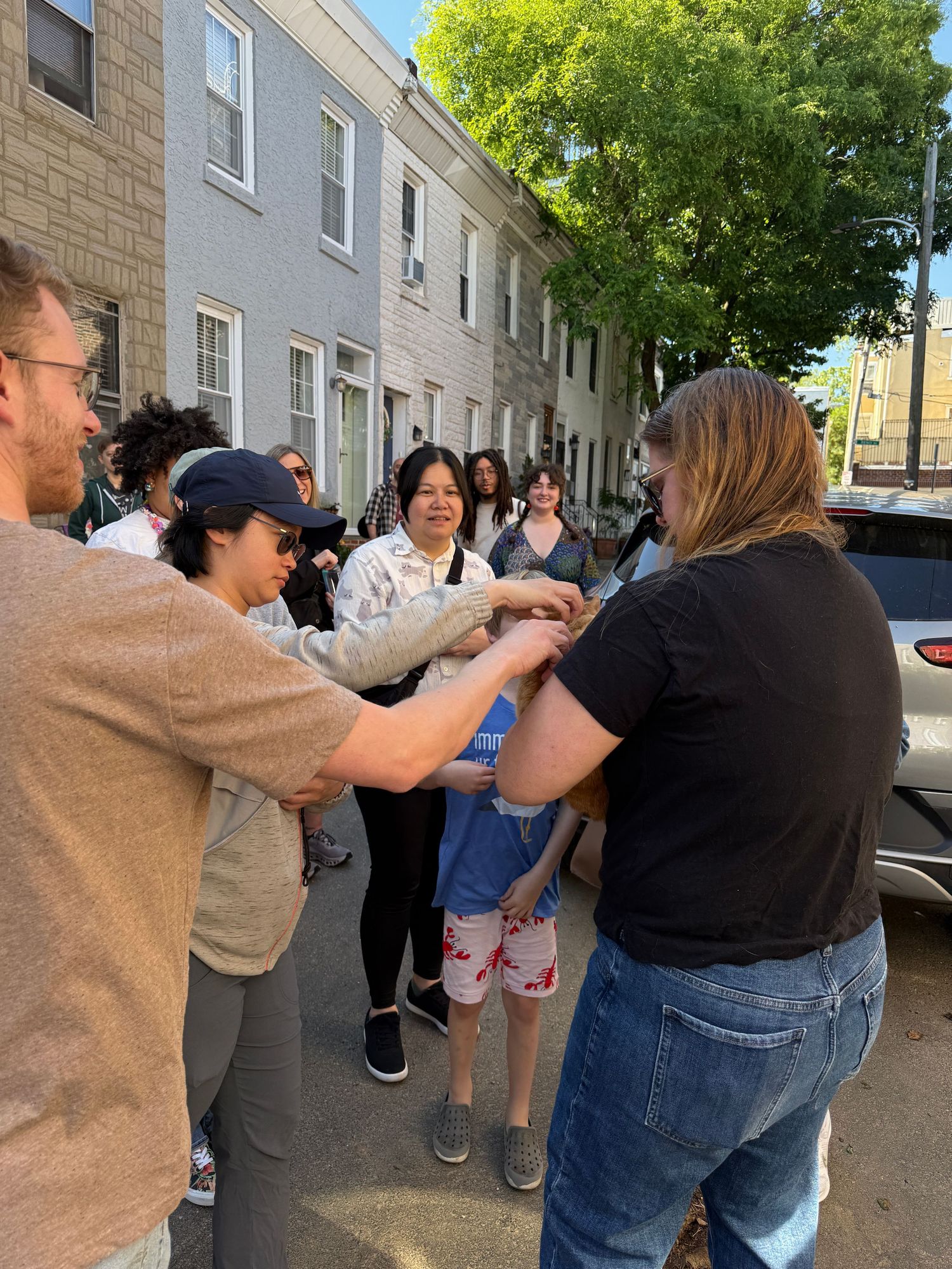 Group of people gathered on a city street, socializing and shaking hands on a sunny day near rowhouses.