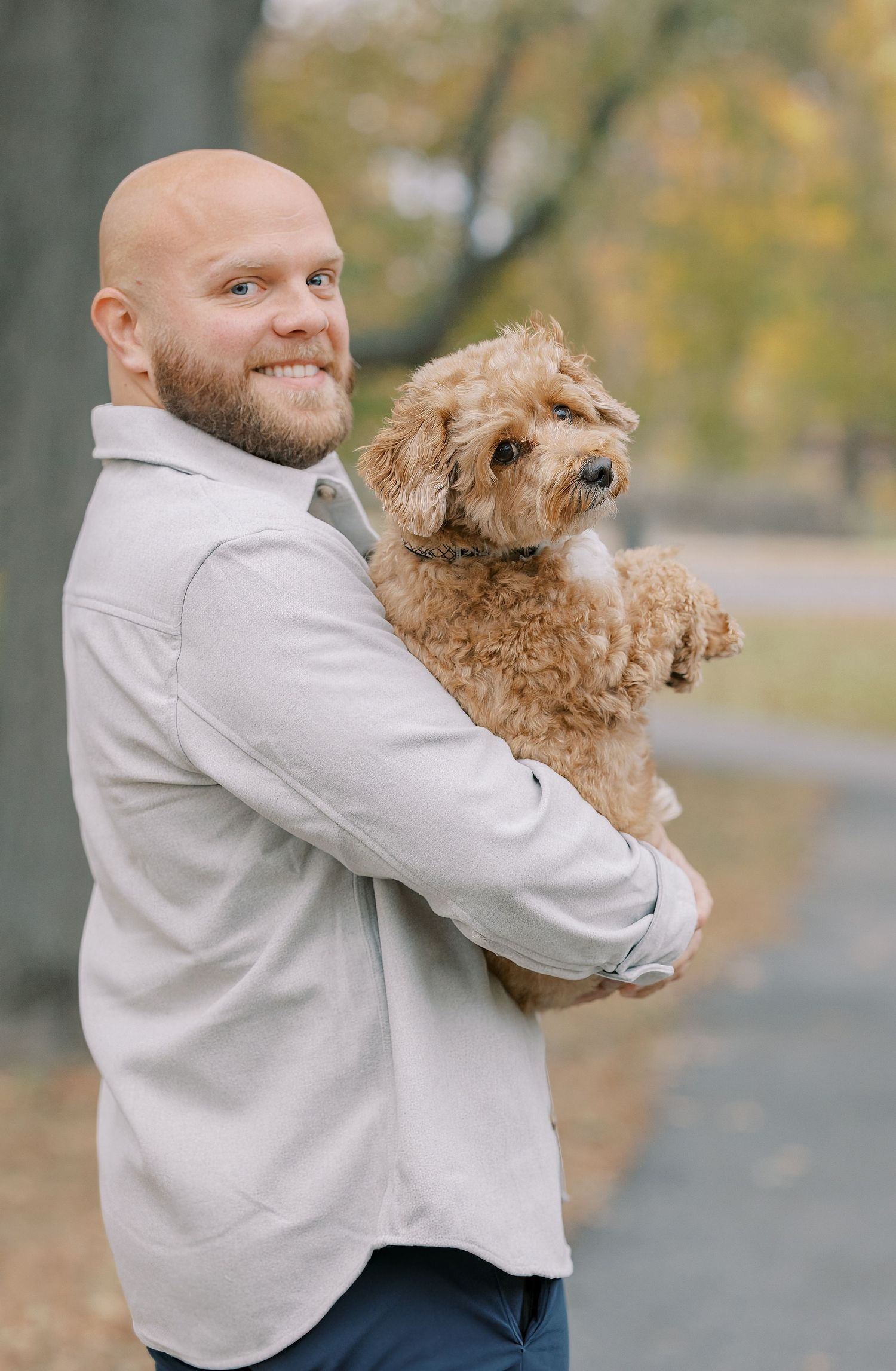 A person in a light gray sweater hugs and plays with their fluffy goldendoodle dog in a park during autumn.
