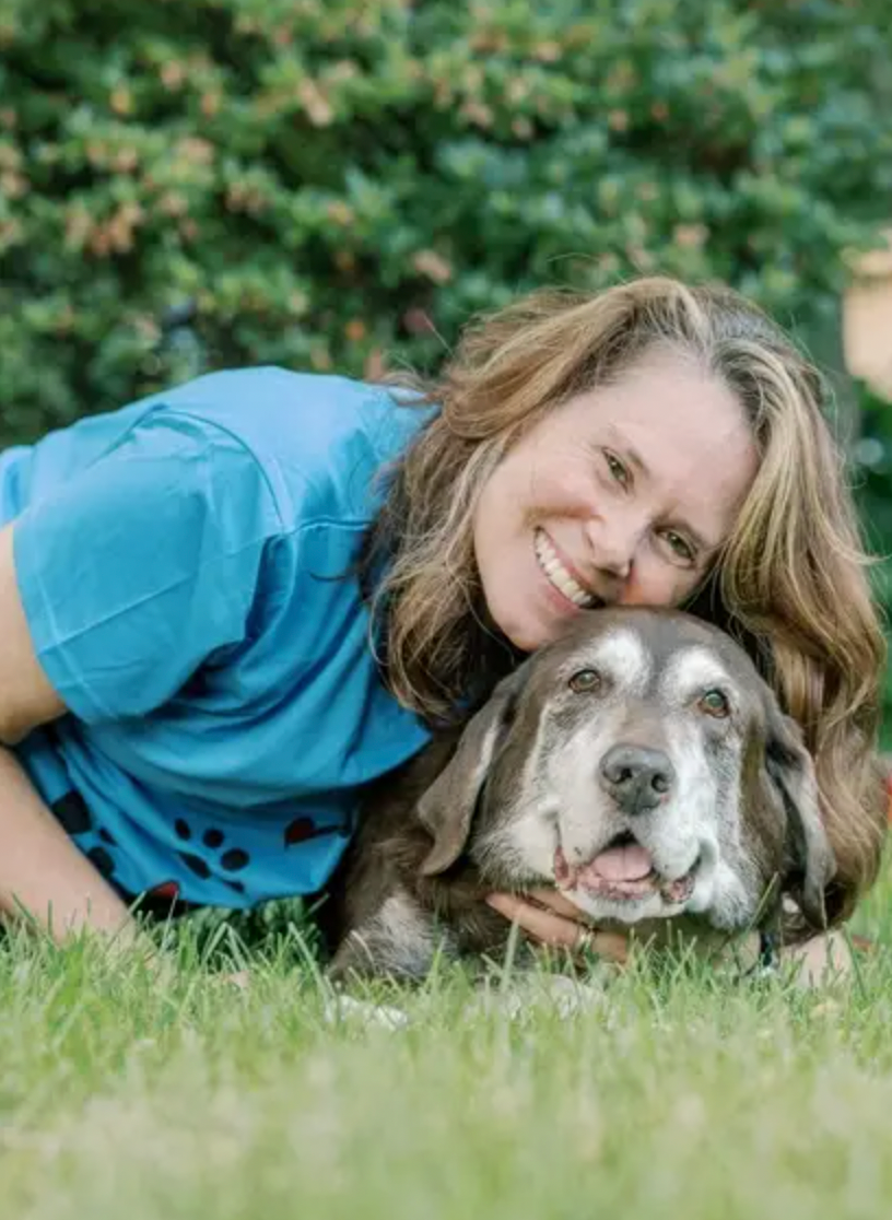 A person in a blue shirt lies in green grass next to a happy senior gray dog in a heartwarming outdoor scene.
