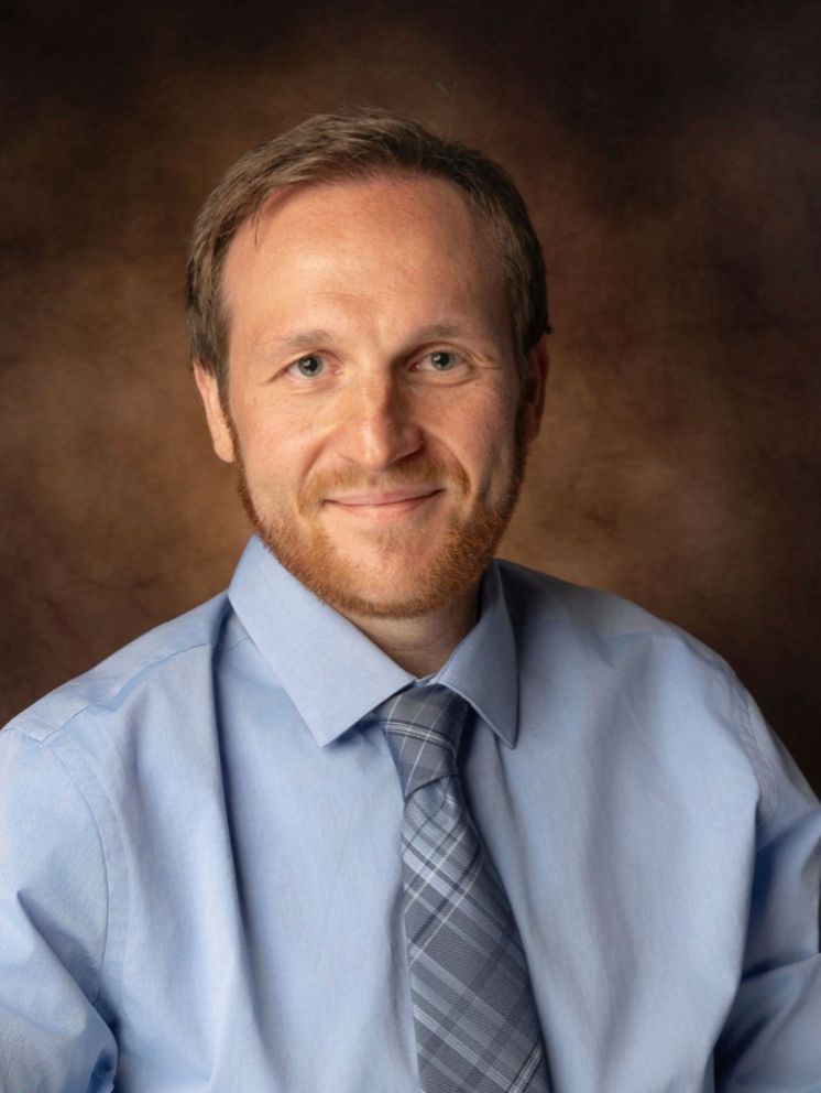 Professional headshot of a smiling man with red beard wearing a light blue shirt and plaid tie against brown background.