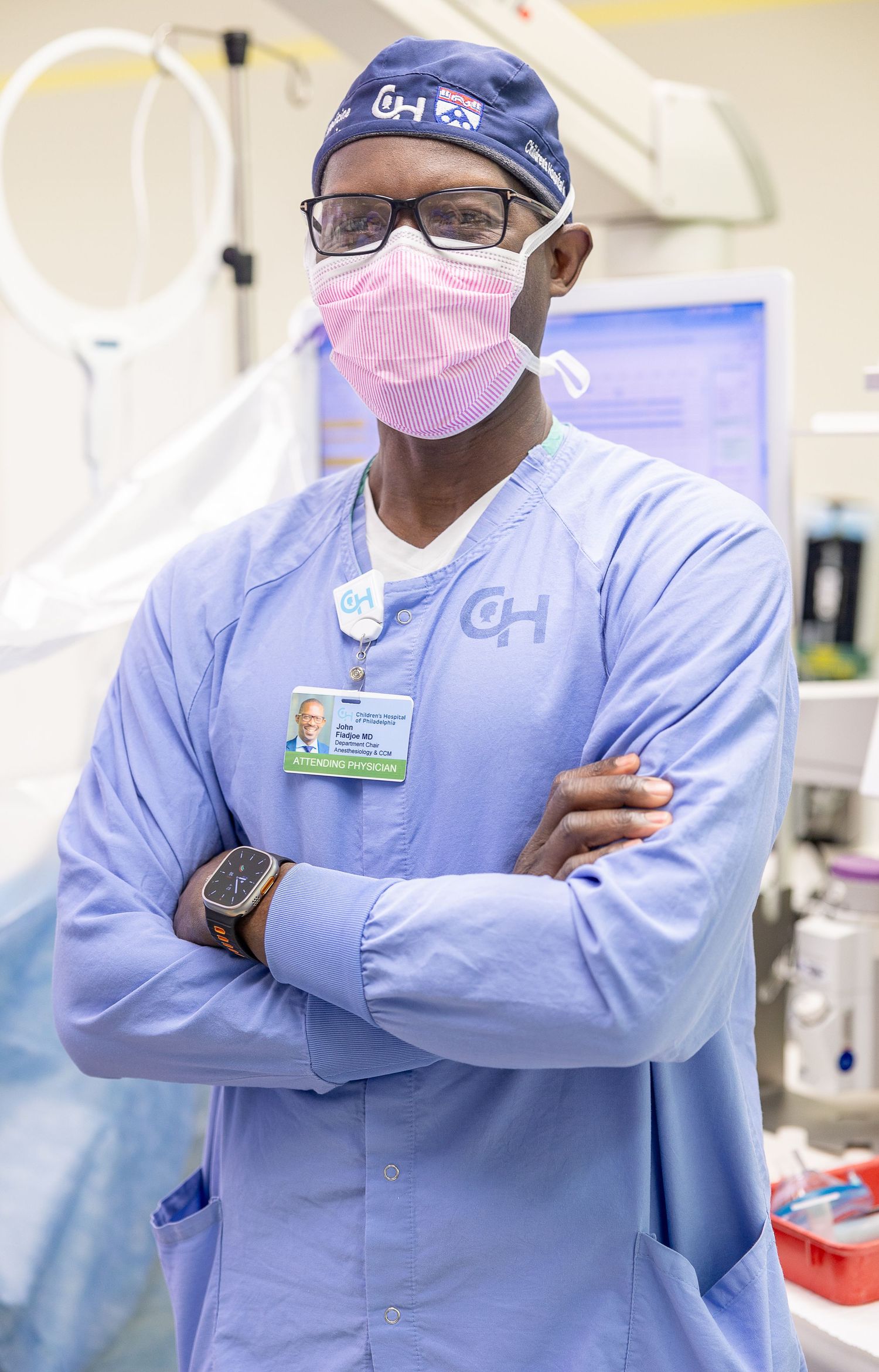 Healthcare worker in light blue scrubs and pink face mask stands with arms crossed in medical setting.