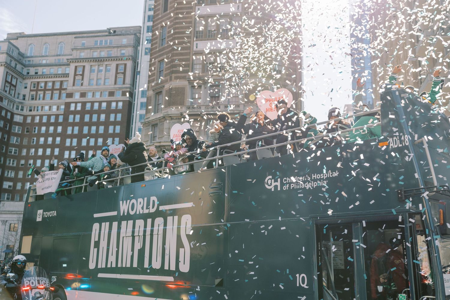 Confetti rains down during Eagles Super Bowl parade on city street with tall buildings.