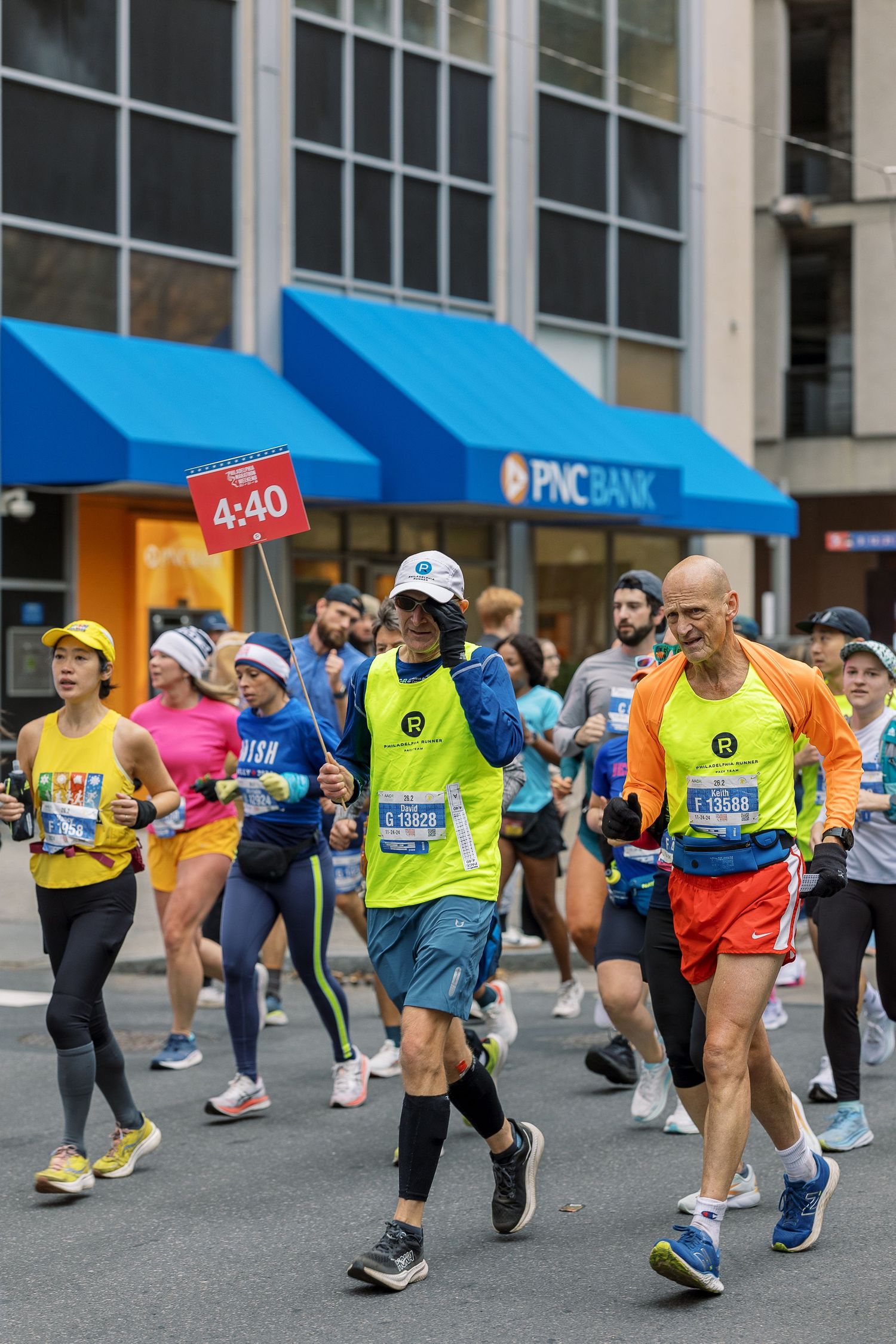 Marathon runners participate in a race passing by PNC Bank with blue awnings and race time markers.