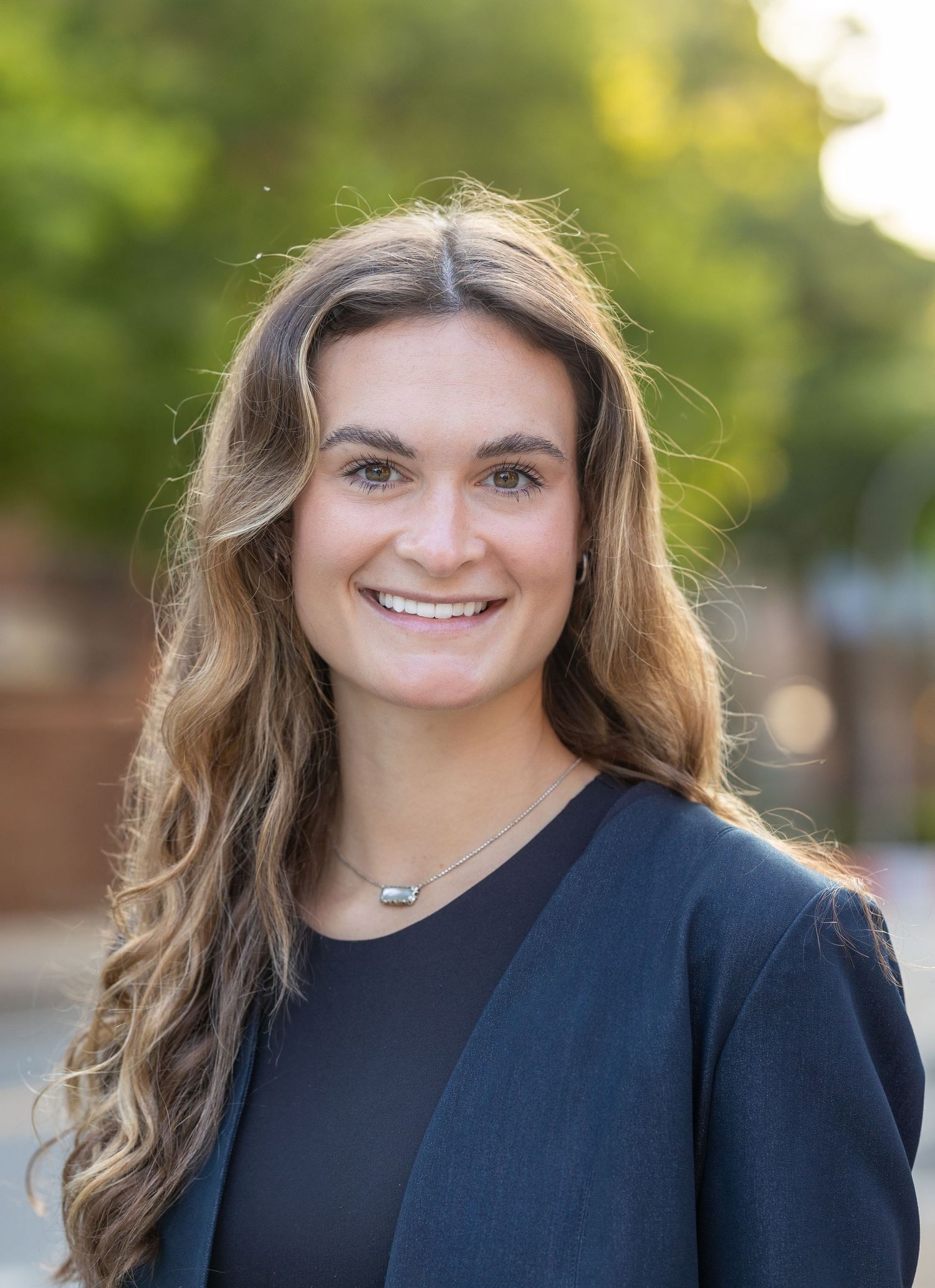 Professional headshot of someone in business attire with wavy hair against blurred outdoor background.