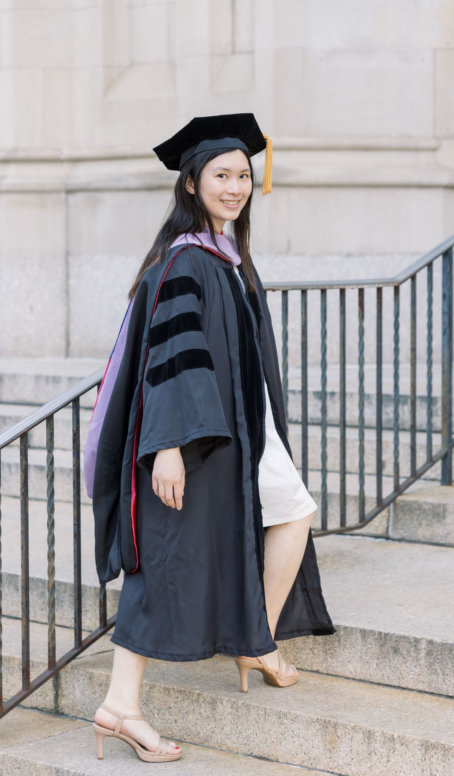 Young woman in doctoral graduation cap and gown poses on stone steps, smiling in heels.