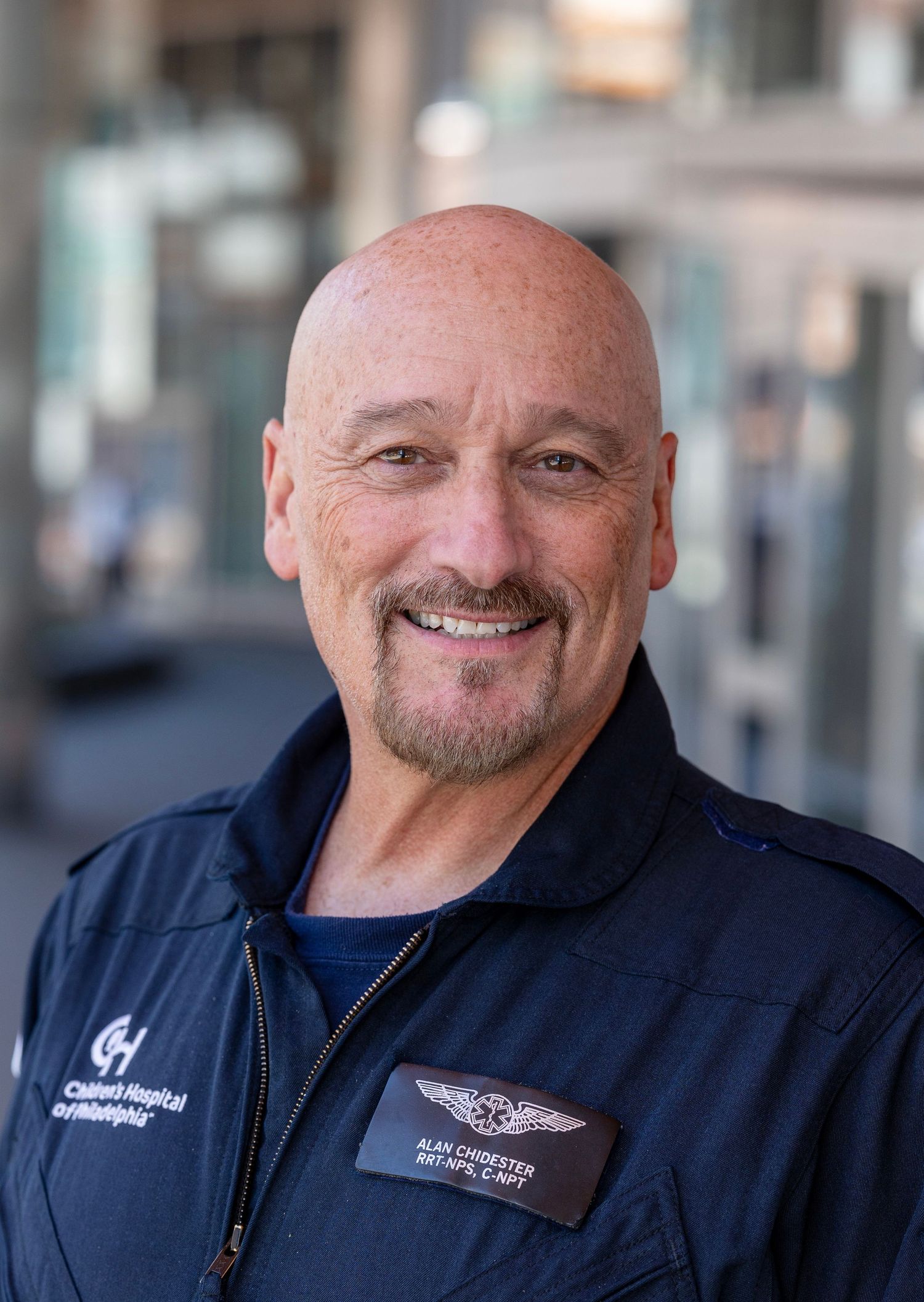 Staff member in a navy uniform jacket displays a friendly smile against a blurred background.