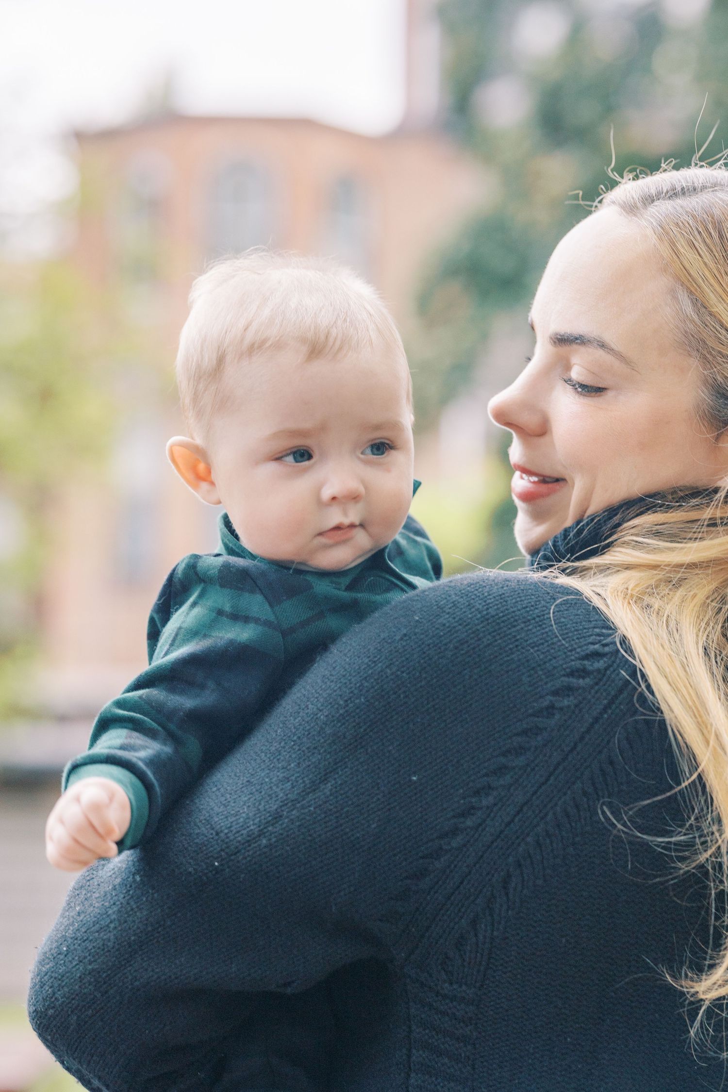 Baby wearing dark sweater being held against caregiver's shoulder in outdoor setting.