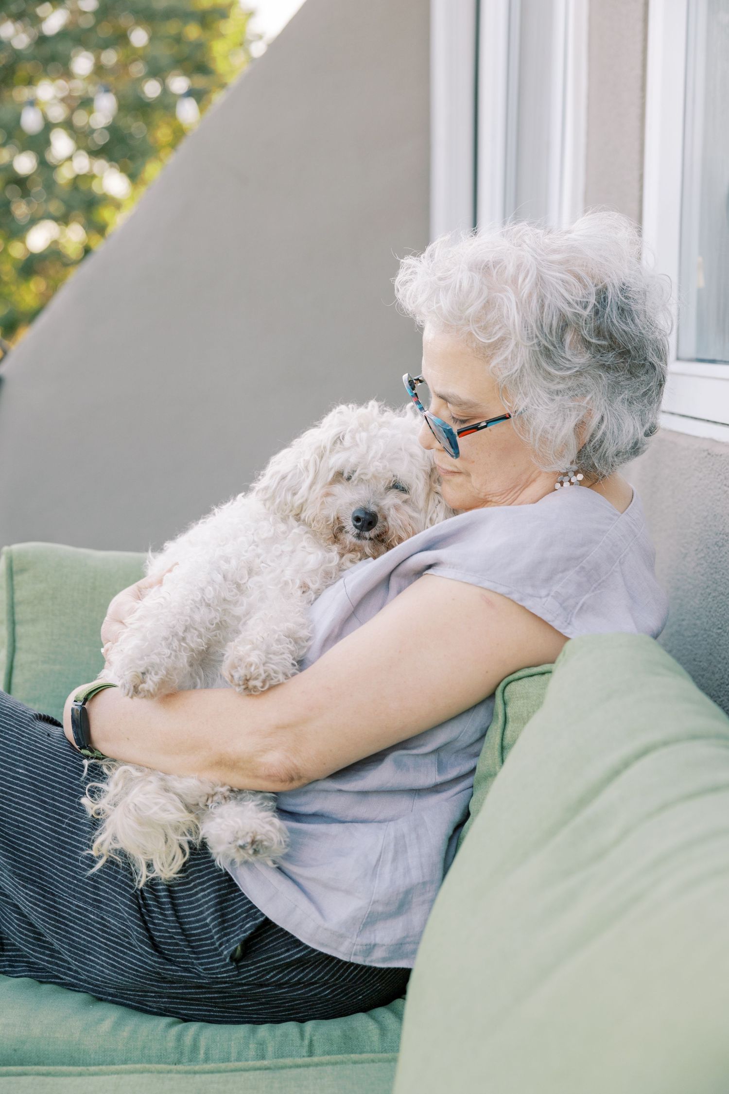 A small white fluffy dog cuddles on a green couch with someone in a gray shirt on a sunny afternoon.