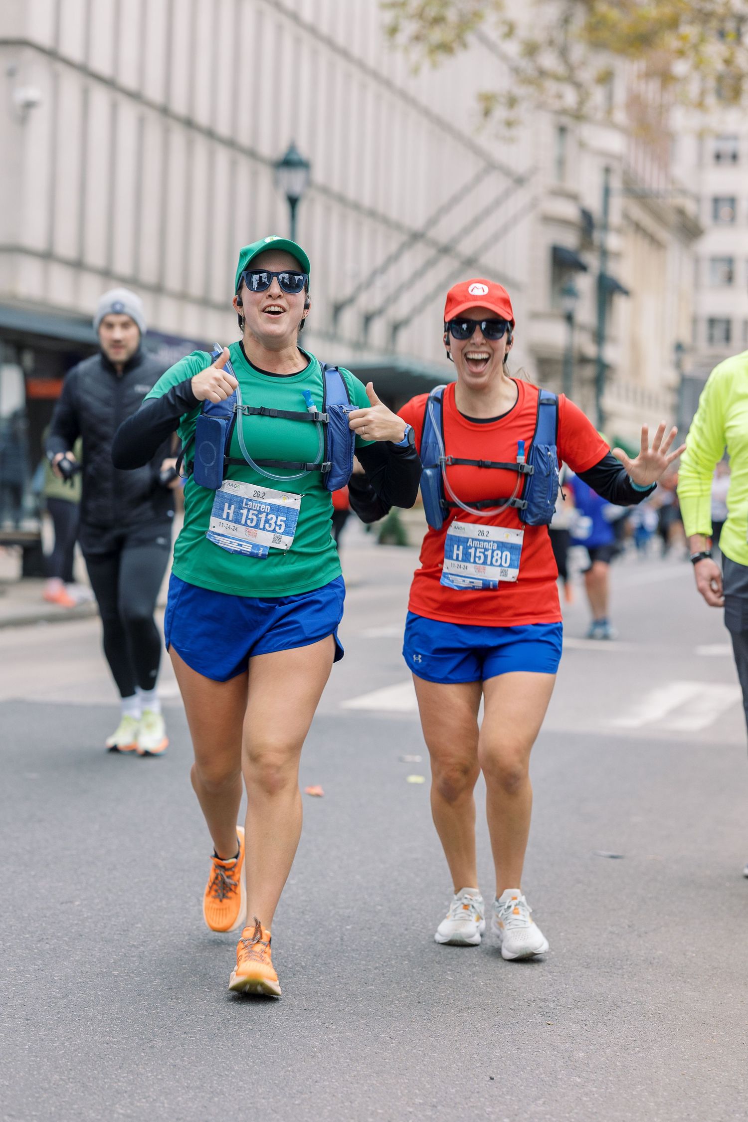 Marathon runners in colorful athletic wear giving thumbs up while running through city streets.