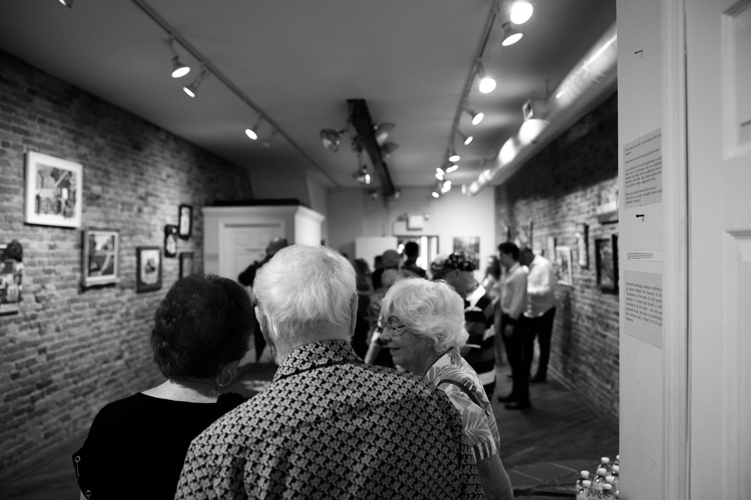 People view artwork in a gallery with exposed brick walls and track lighting.