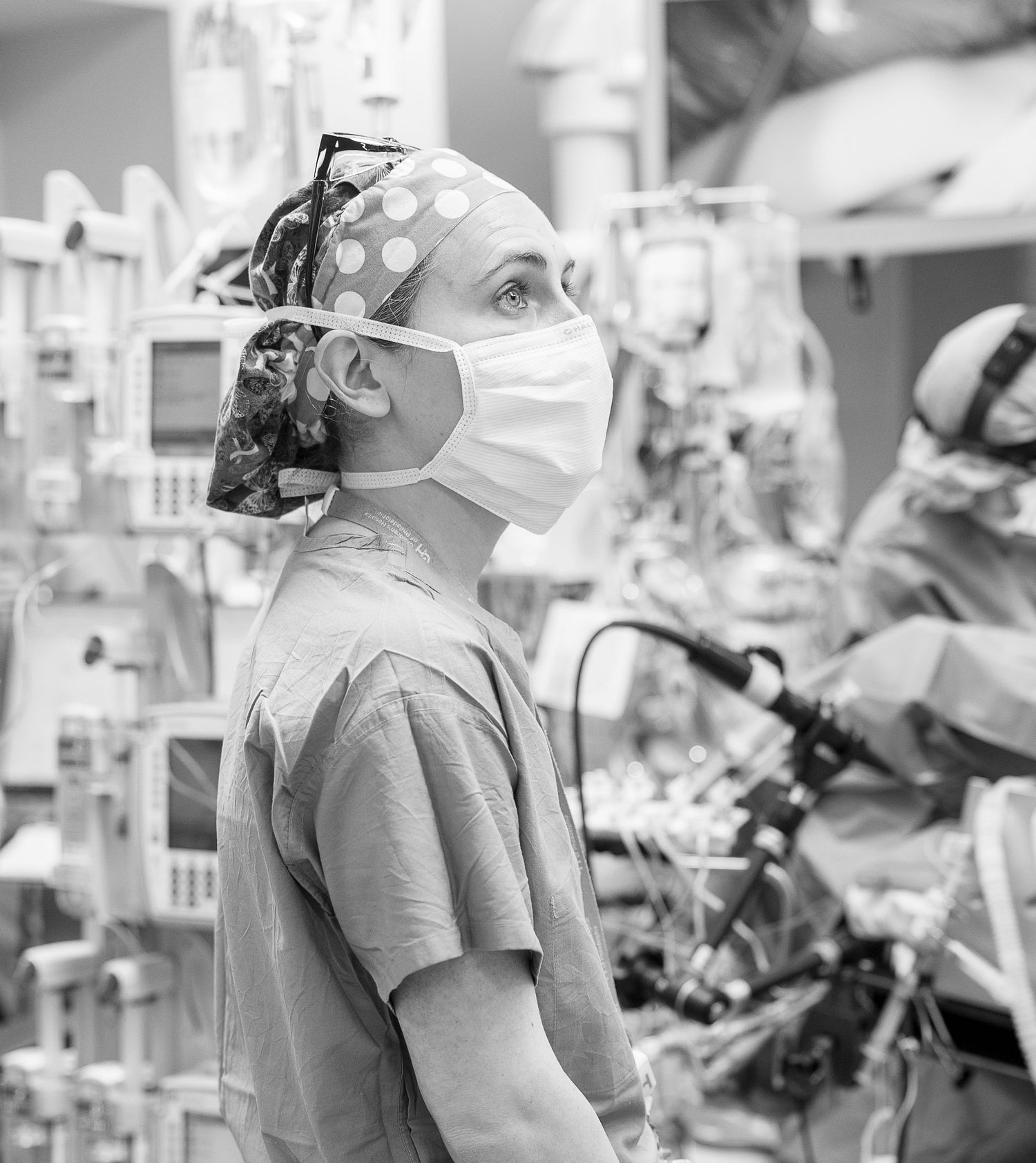 A masked surgeon in scrubs and patterned cap stands alert amid medical equipment in a hospital operating room.