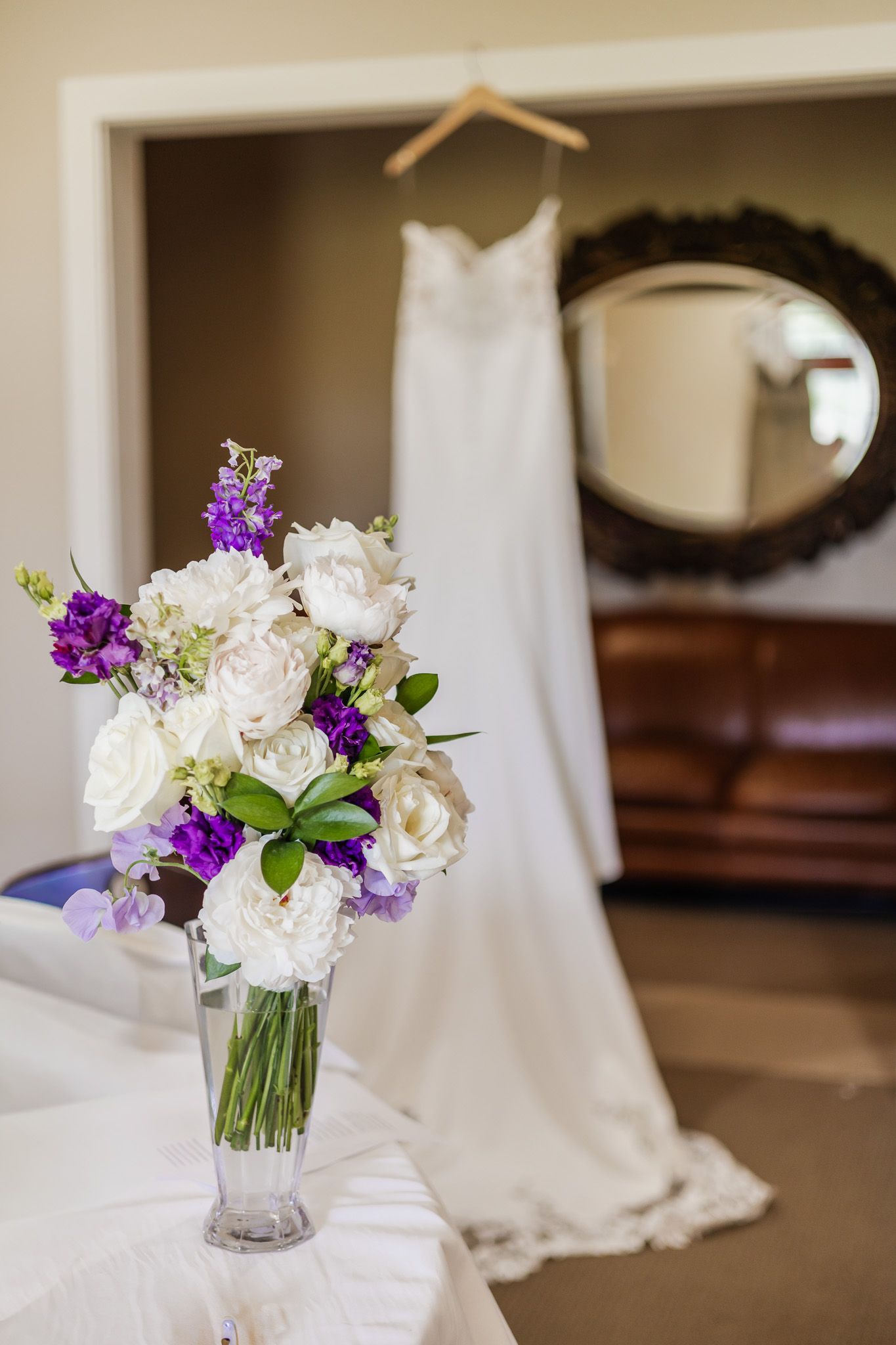White and purple wedding bouquet in glass vase with bridal gown hanging in background near ornate mirror.