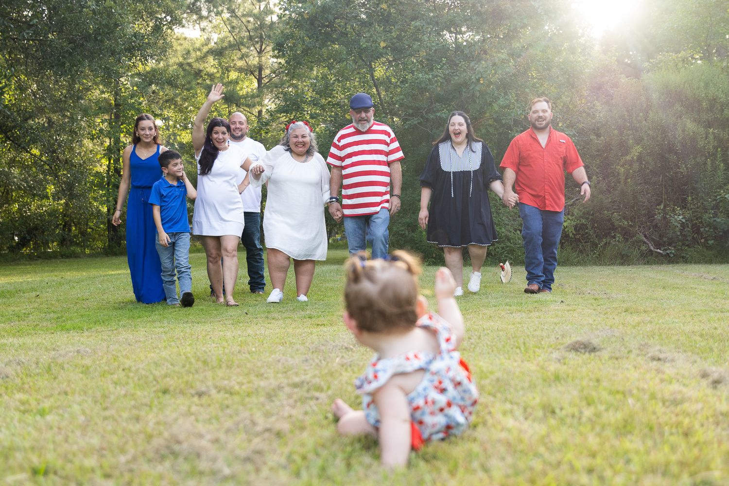 A family group walks across a green lawn toward a baby sitting in the foreground during an outdoor photo session.