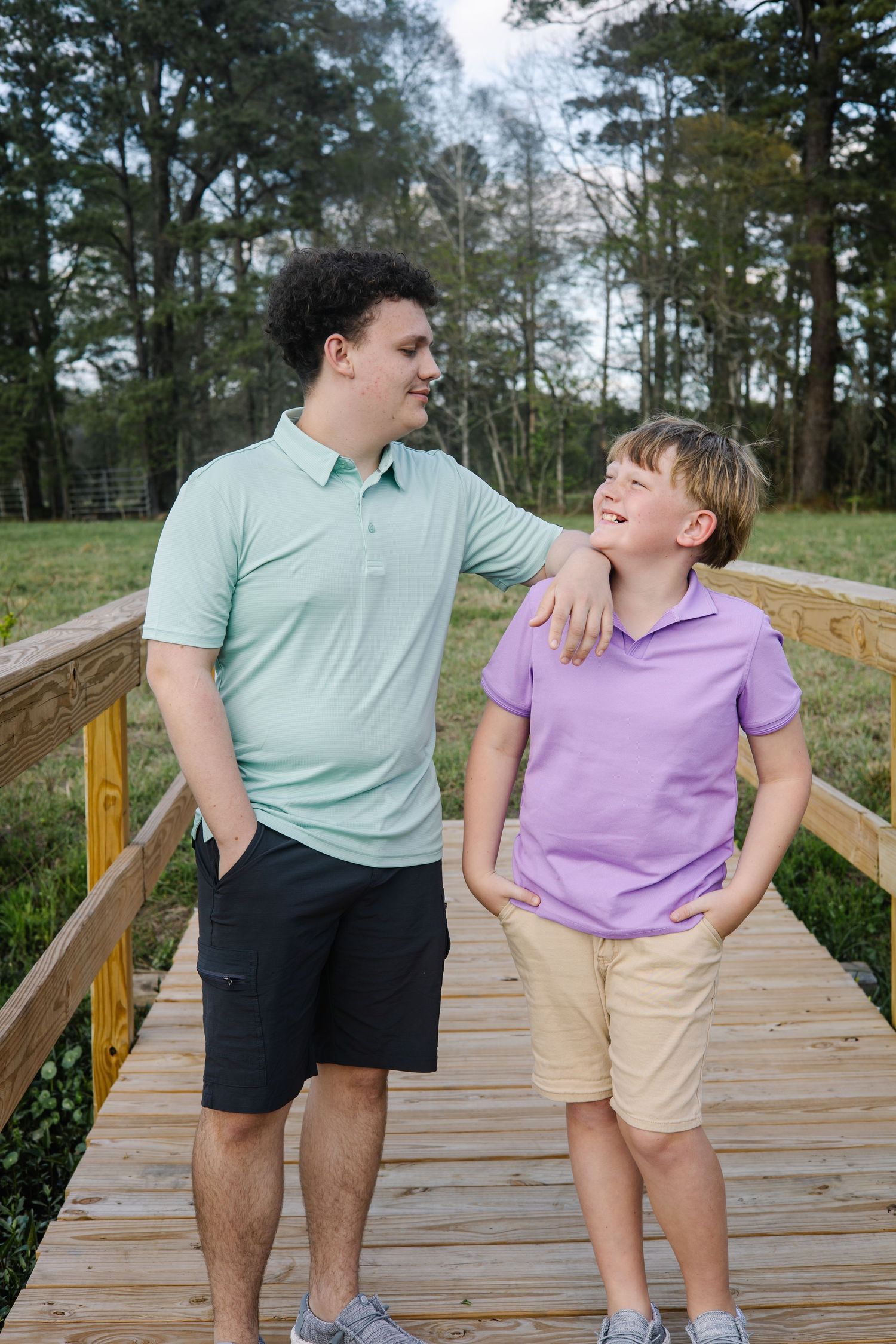 Two boys in pastel polo shirts share a friendly moment on a wooden bridge surrounded by trees.