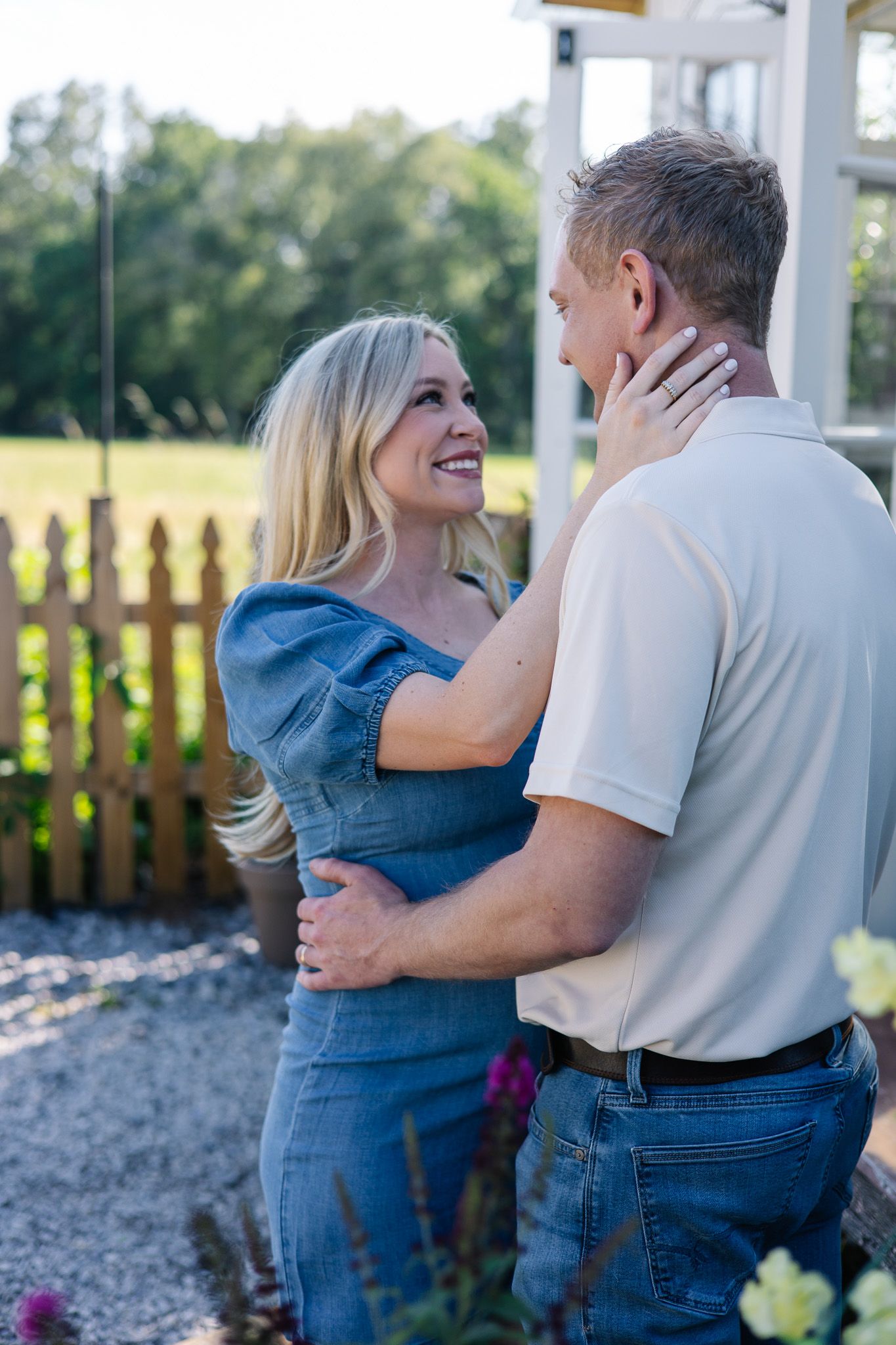 Smiling blonde woman in denim dress gazing lovingly at her partner while holding his face in a sunny garden.