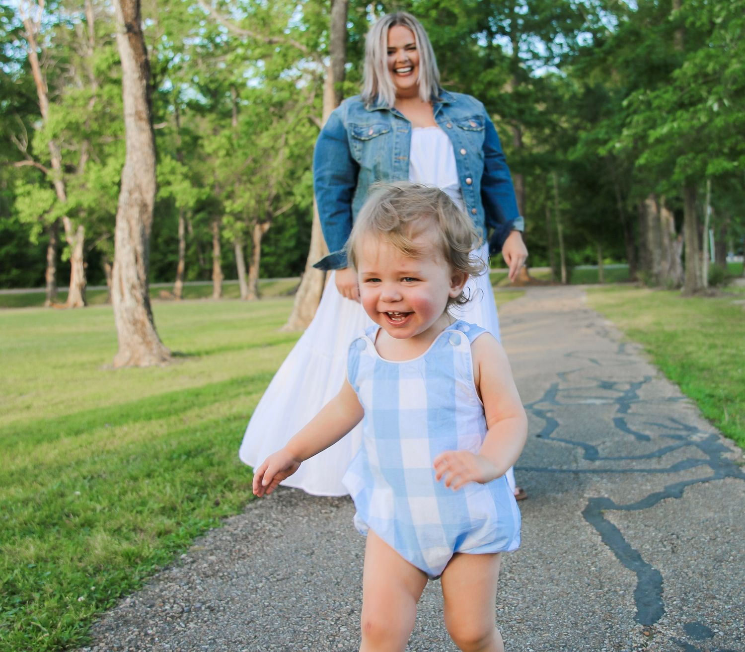 Laughing toddler in blue gingham romper running on a tree-lined path with mother behind.