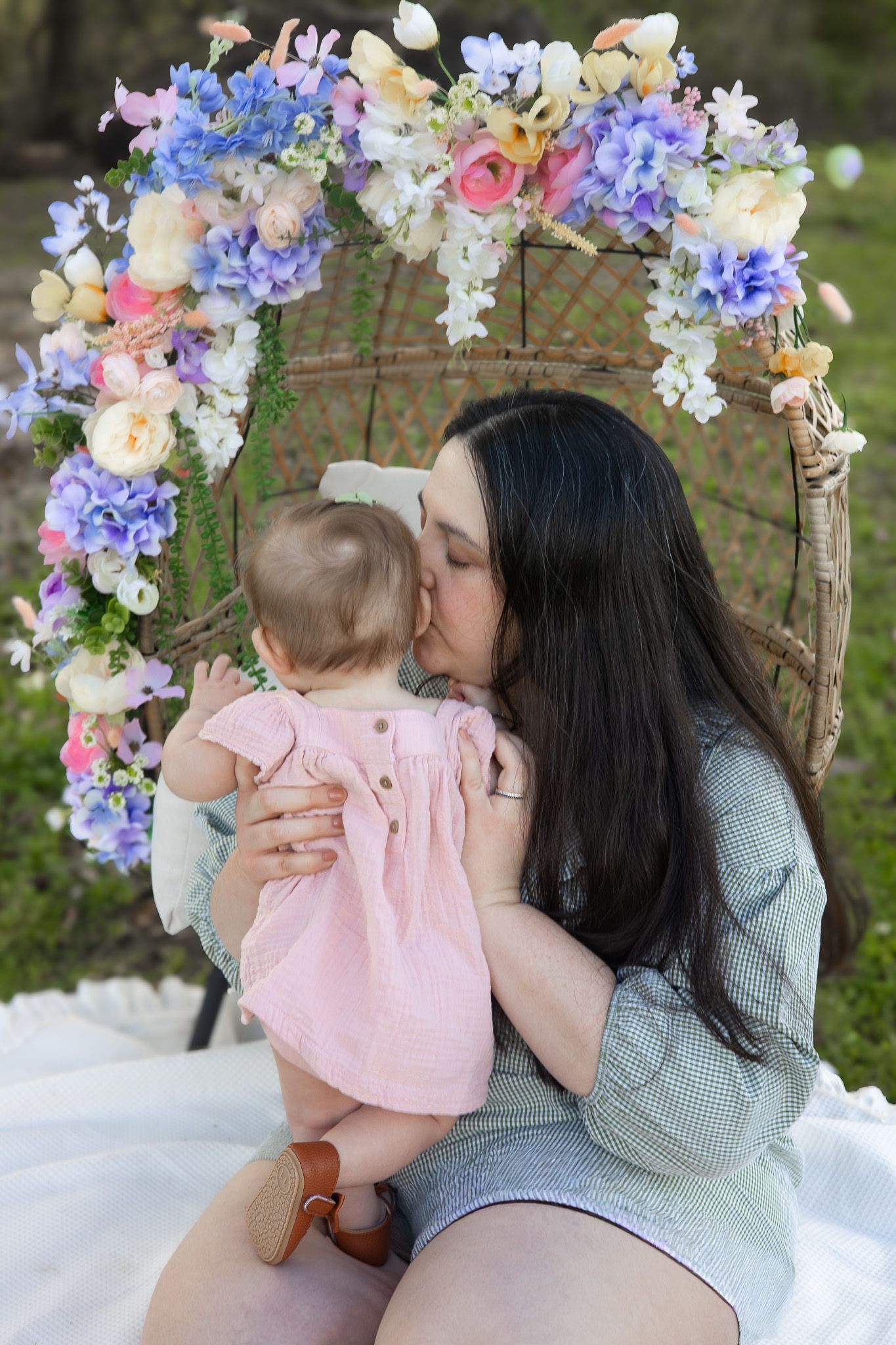 A mother tenderly kisses her baby girl under a beautiful floral arch in an outdoor setting.