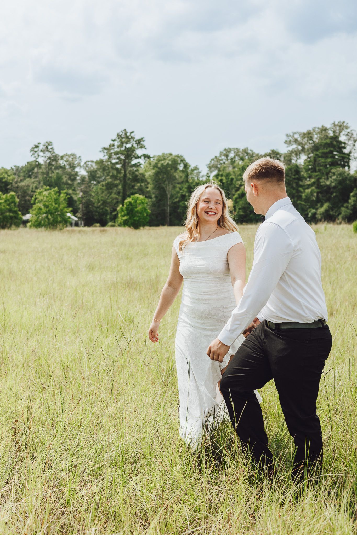A joyful bride in a white dress holds hands with her groom in a sunlit green field, laughing as they walk together.