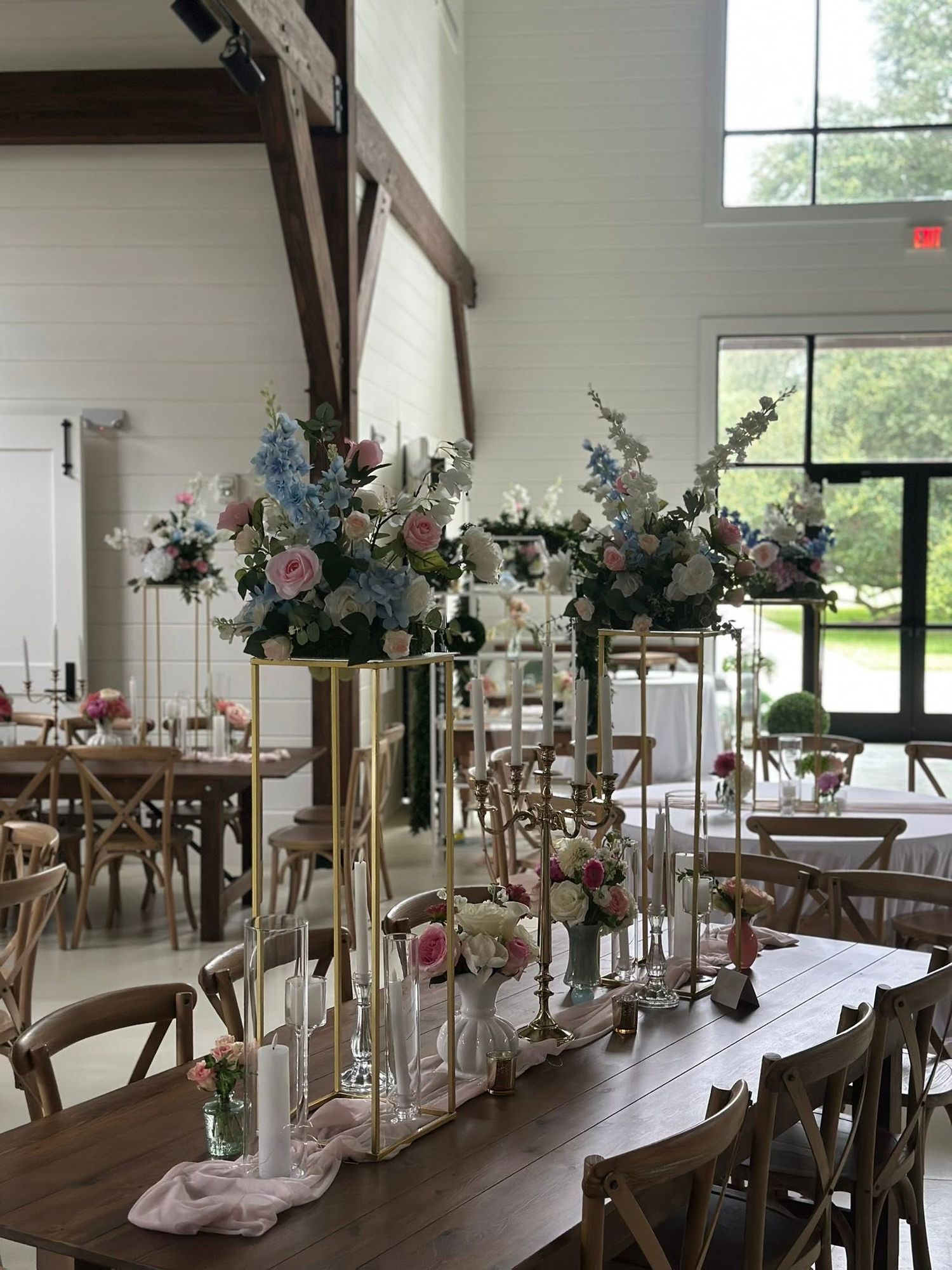 Elegant wedding reception hall with tall gold floral centerpieces featuring pink and blue blooms on rustic wooden farm tables.
