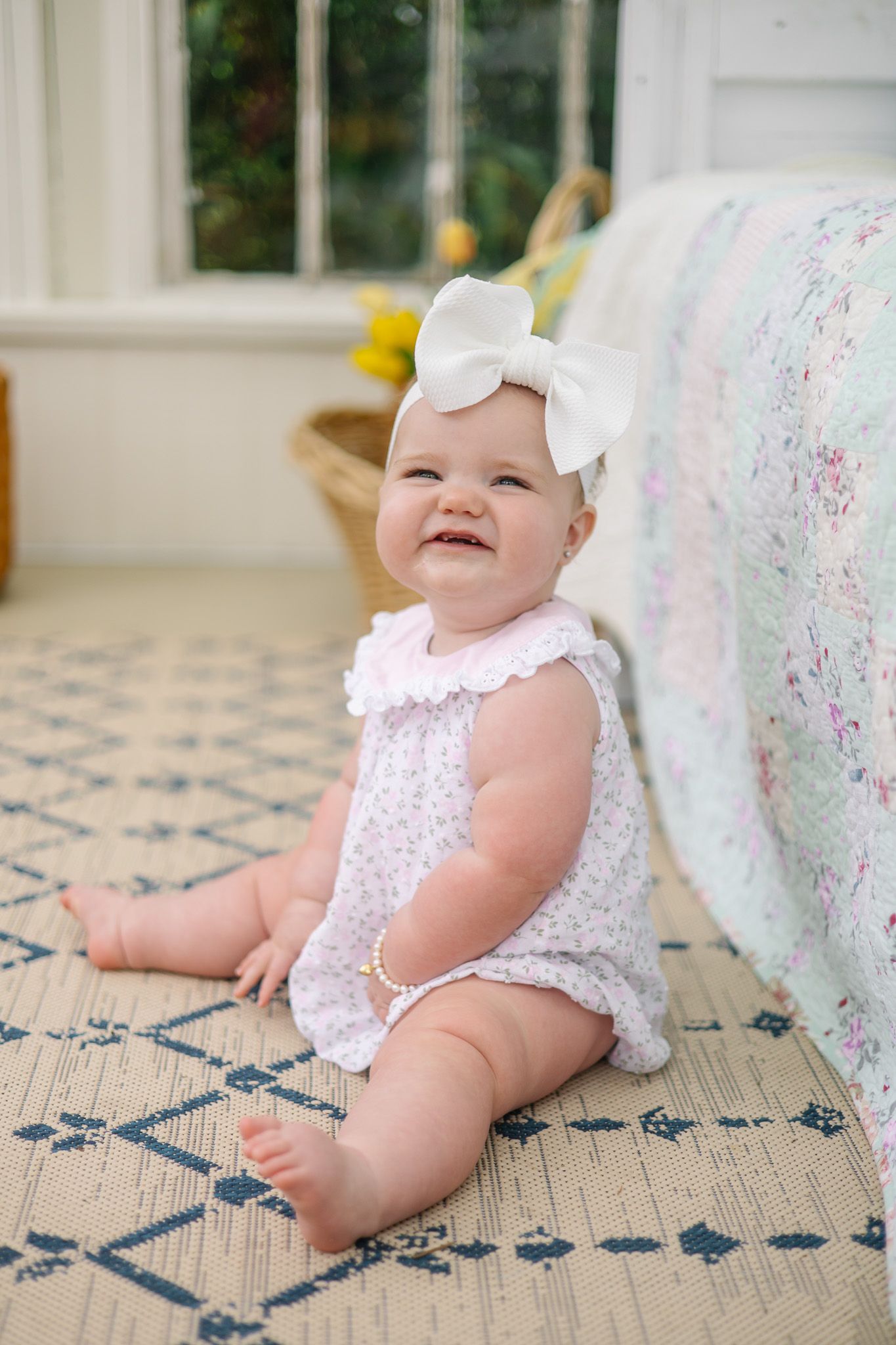 Happy baby girl in pink floral romper with white bow headband sits on patterned rug.