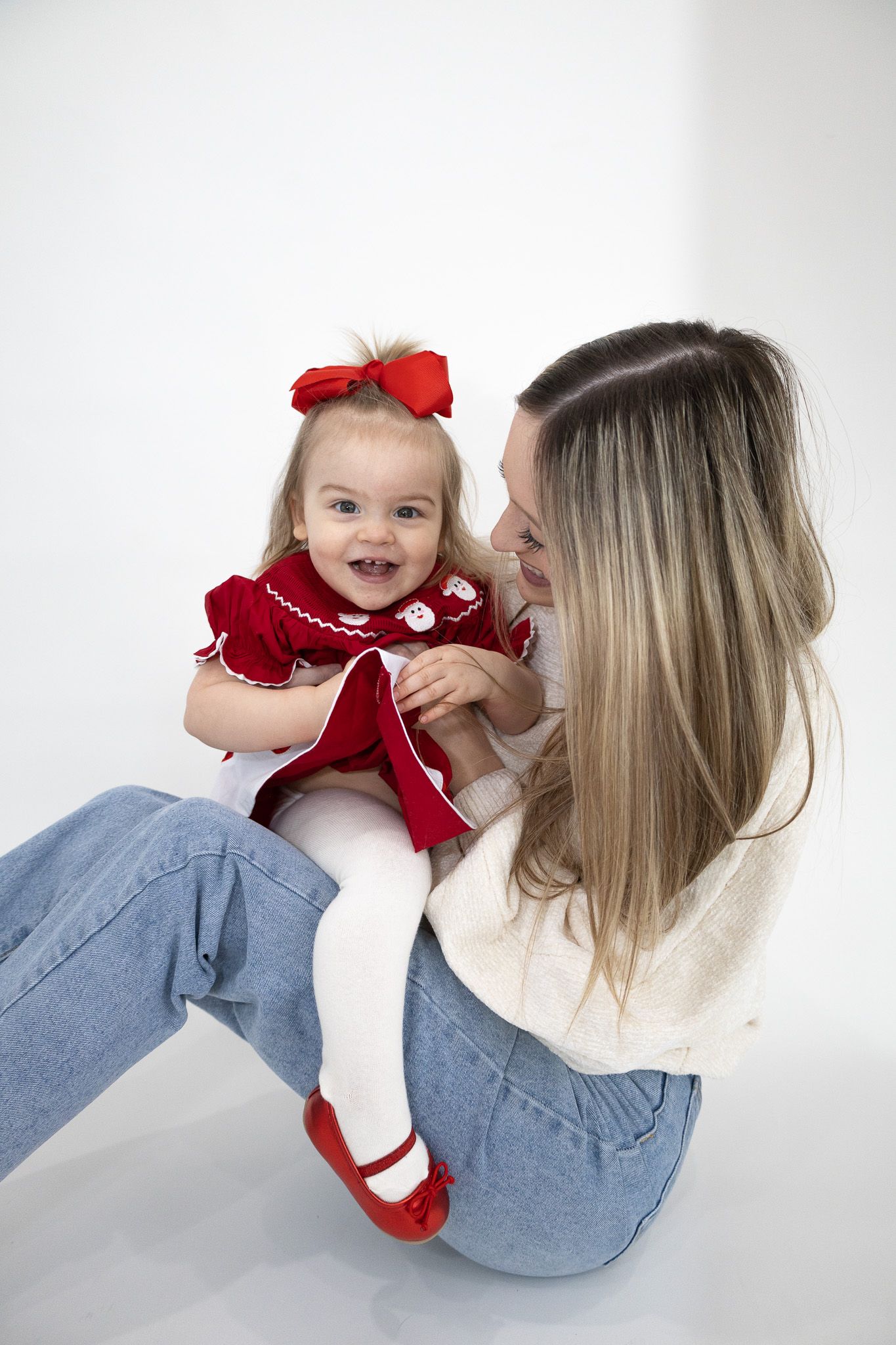 A happy baby girl in a red dress and bow is held by her smiling blonde mother indoors.