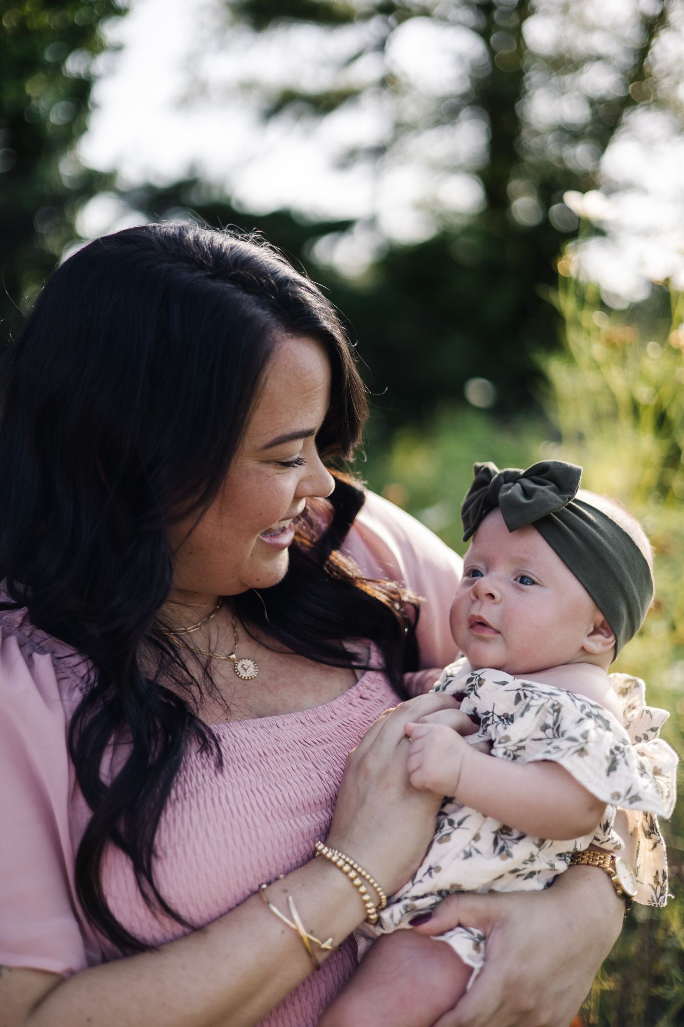 A mother in pink holds her newborn baby girl wearing a floral outfit and dark bow headband outdoors.