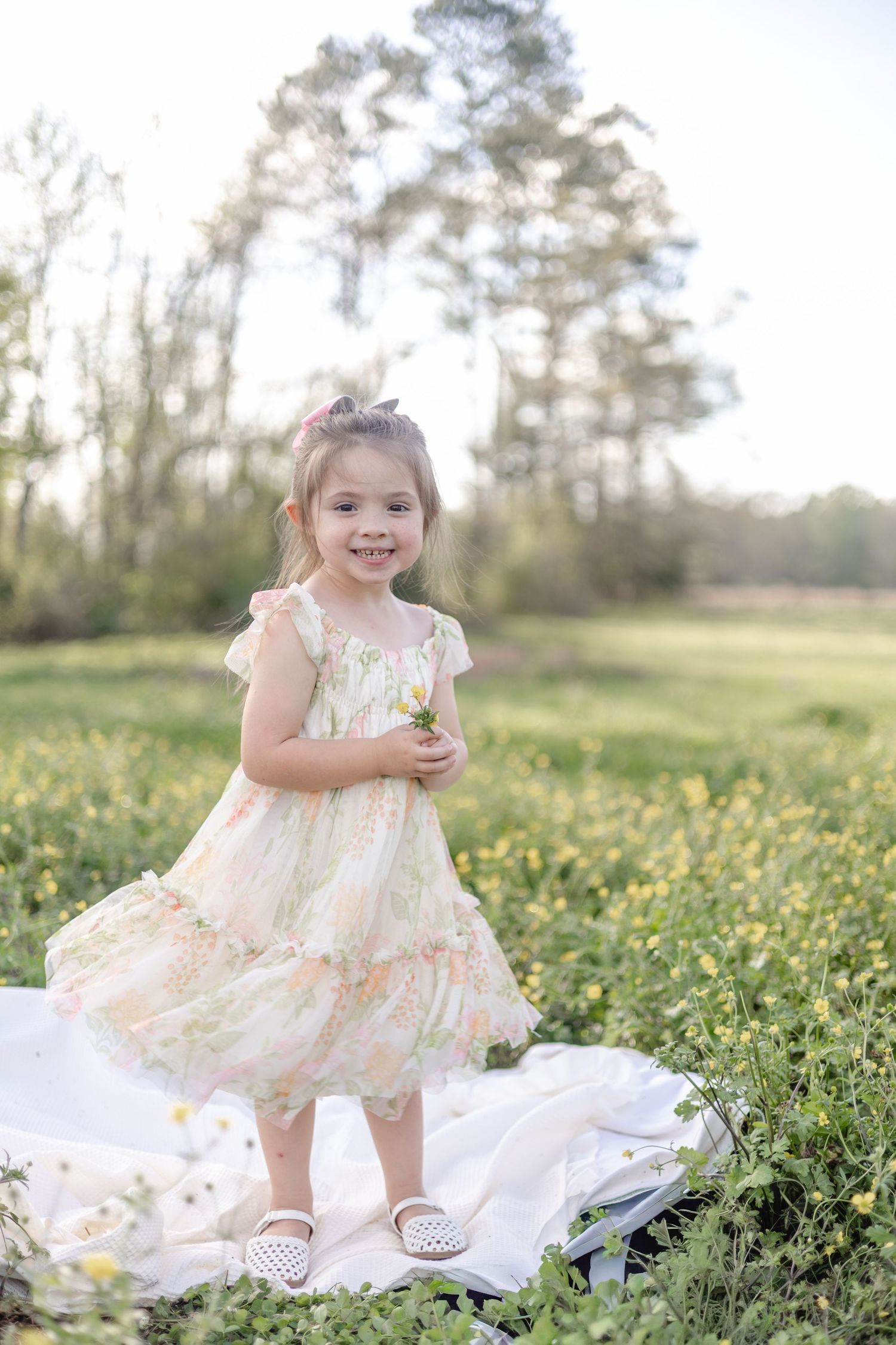 A smiling toddler girl in a floral tulle dress holds wildflowers in a sunny yellow flower field.