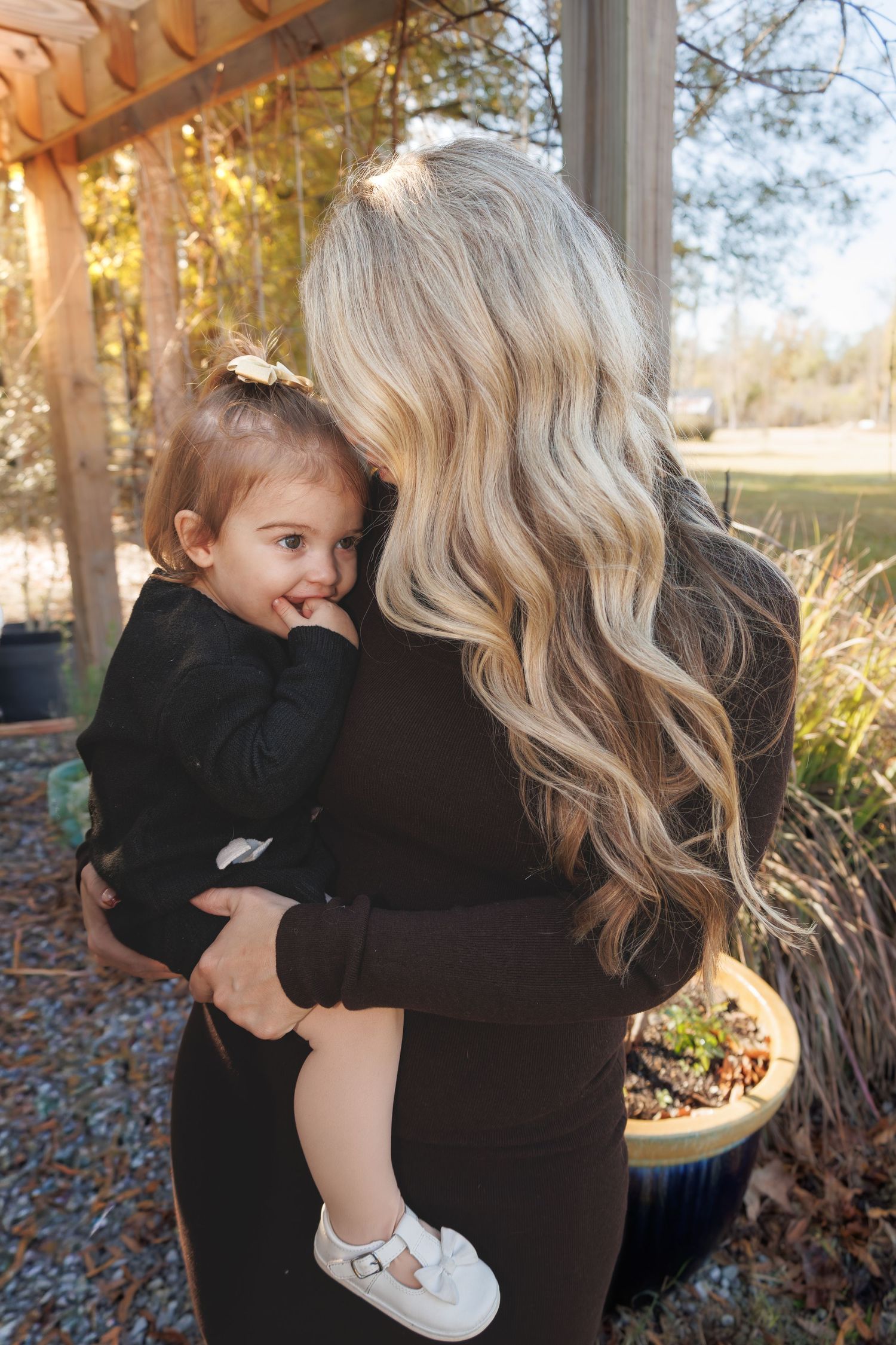 Blonde woman with wavy hair holding a smiling toddler in a black outfit outdoors.