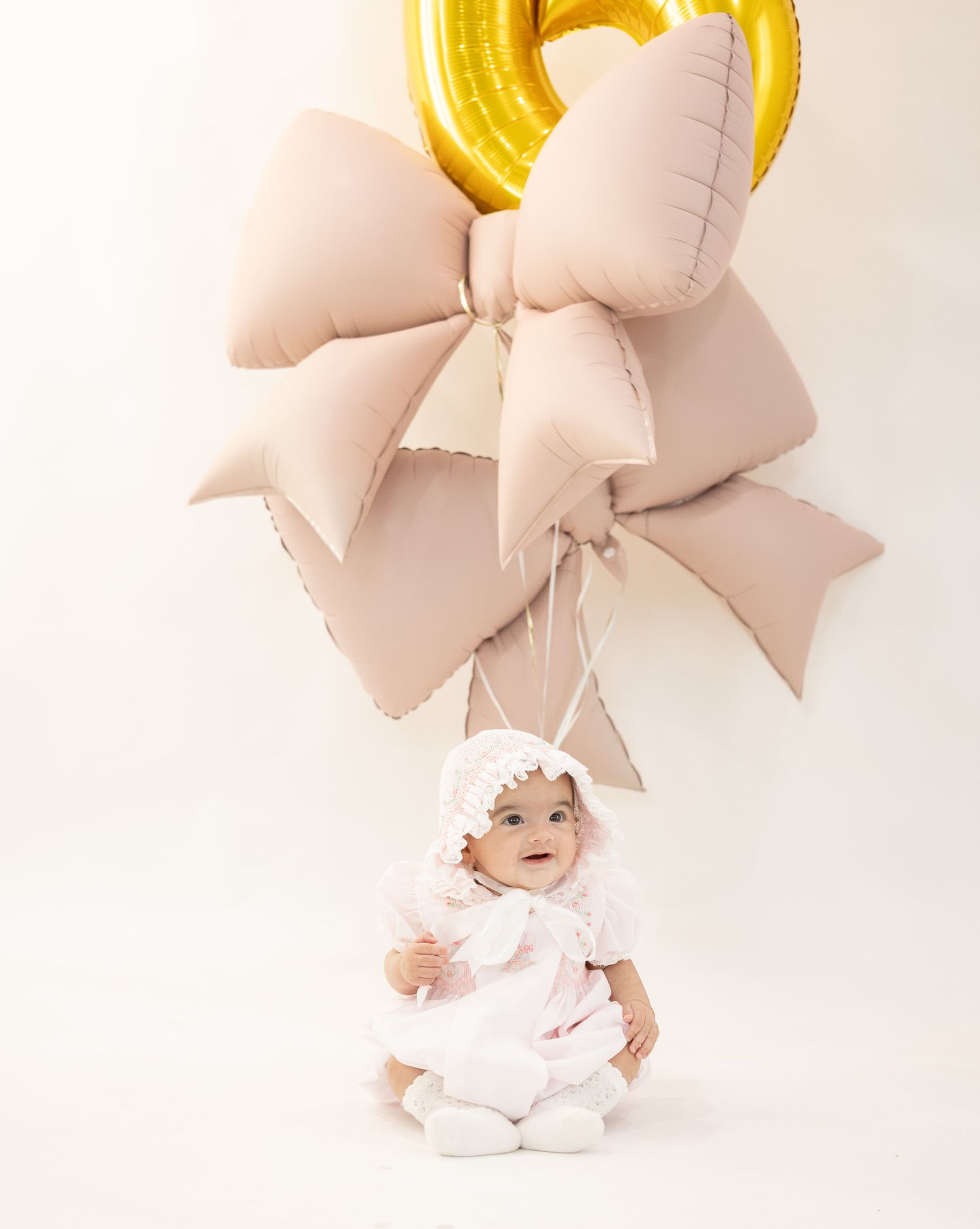 Smiling baby in white dress and bonnet sits before pink bow balloons and gold number balloon.