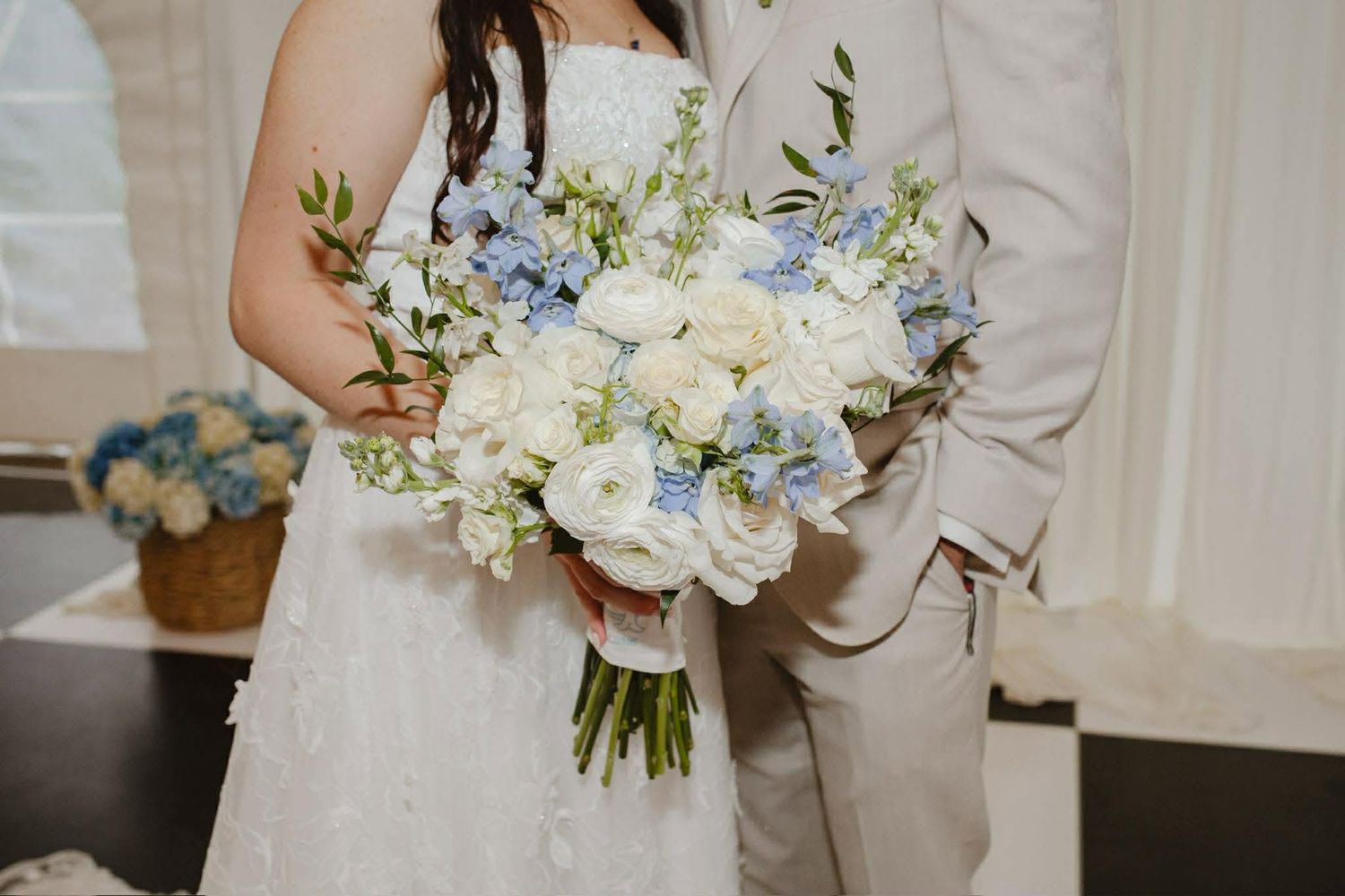 Bride holding a lush wedding bouquet of white ranunculus and blue delphinium flowers, paired with a groom in a tan suit.