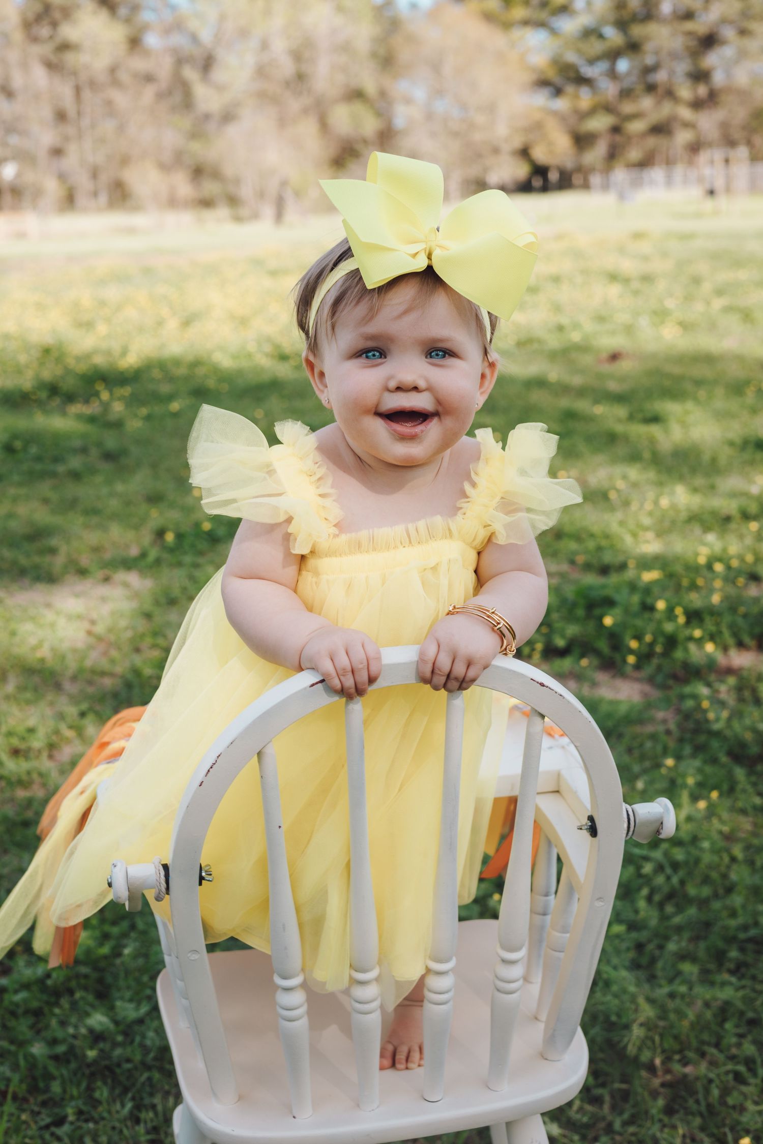 An adorable baby girl in a yellow dress and bow headband stands on a white chair outdoors.