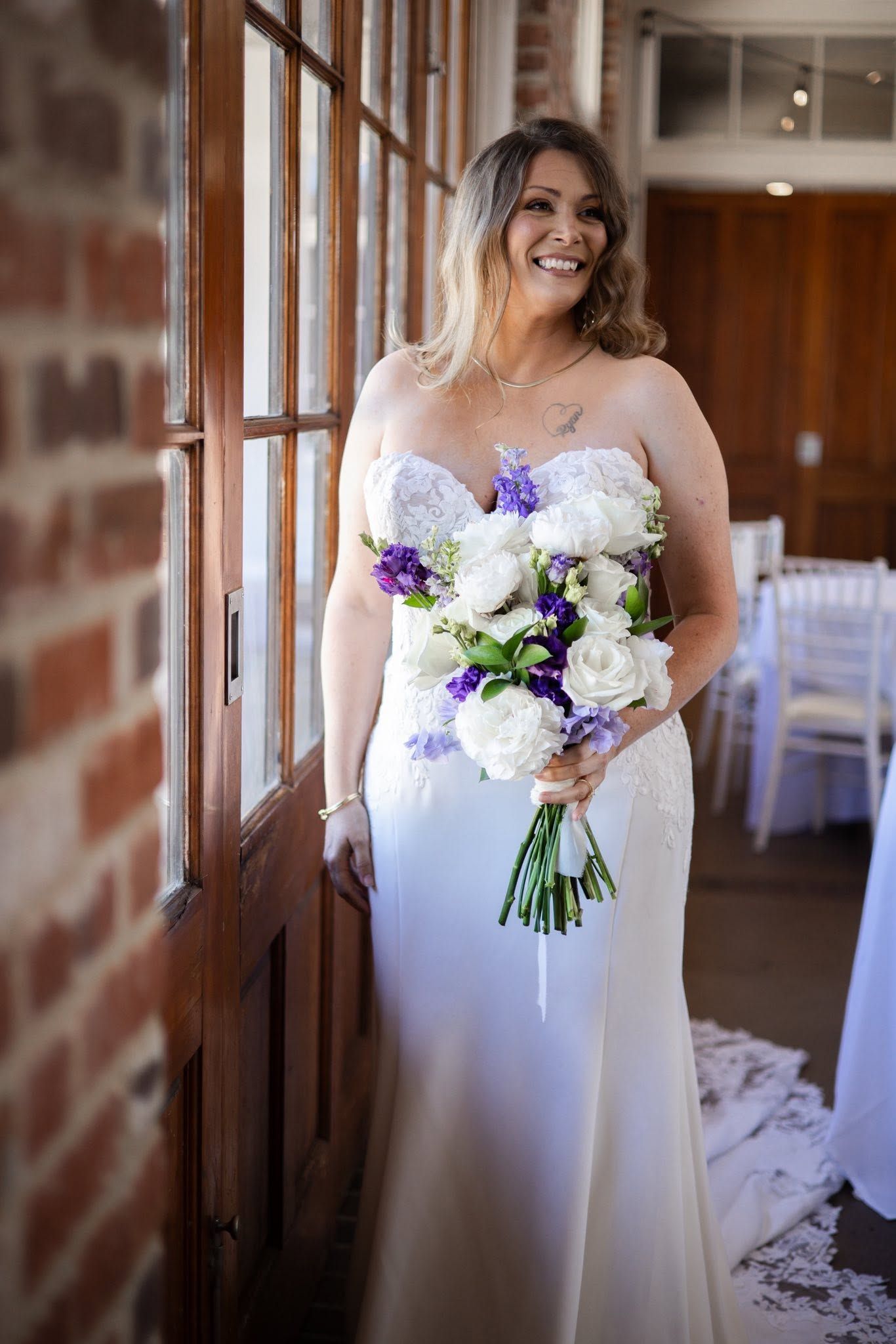 Smiling bride in strapless white gown holding purple and white bouquet, leaning against wooden door in brick venue.