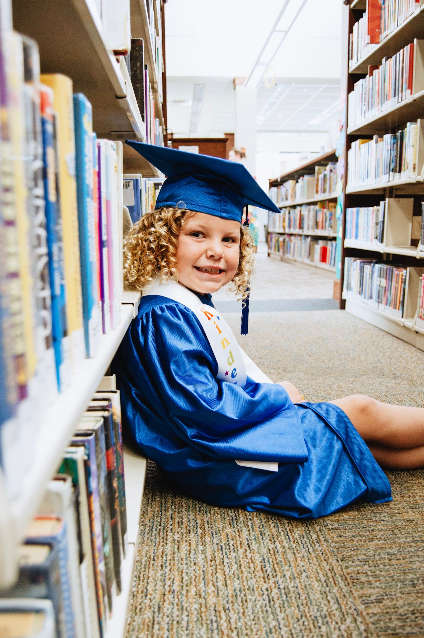 Smiling curly-haired girl in blue graduation cap and gown sitting on library floor beside bookshelves.