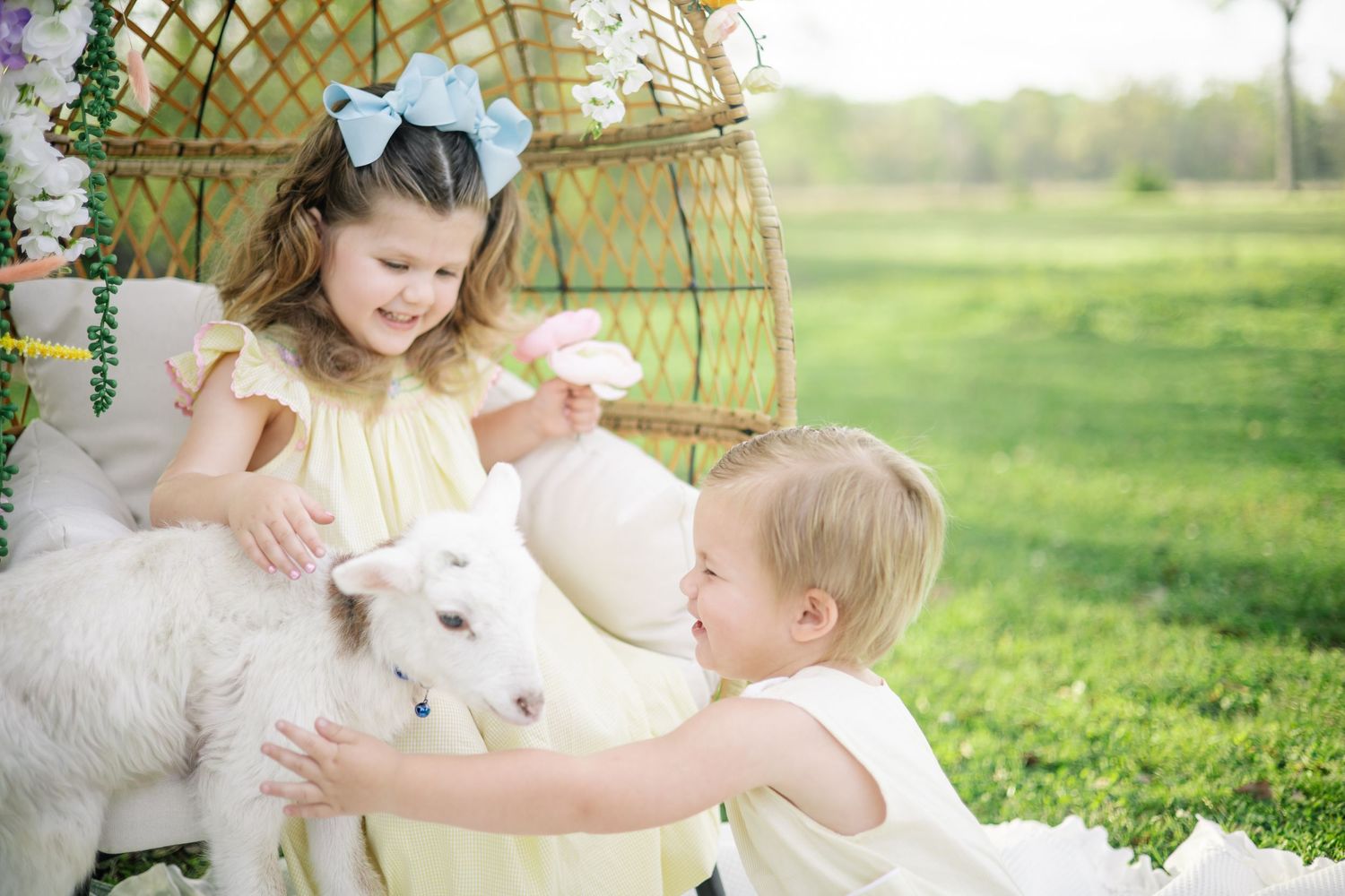 Two young children in yellow outfits petting a white baby goat outdoors near a floral decorated chair.