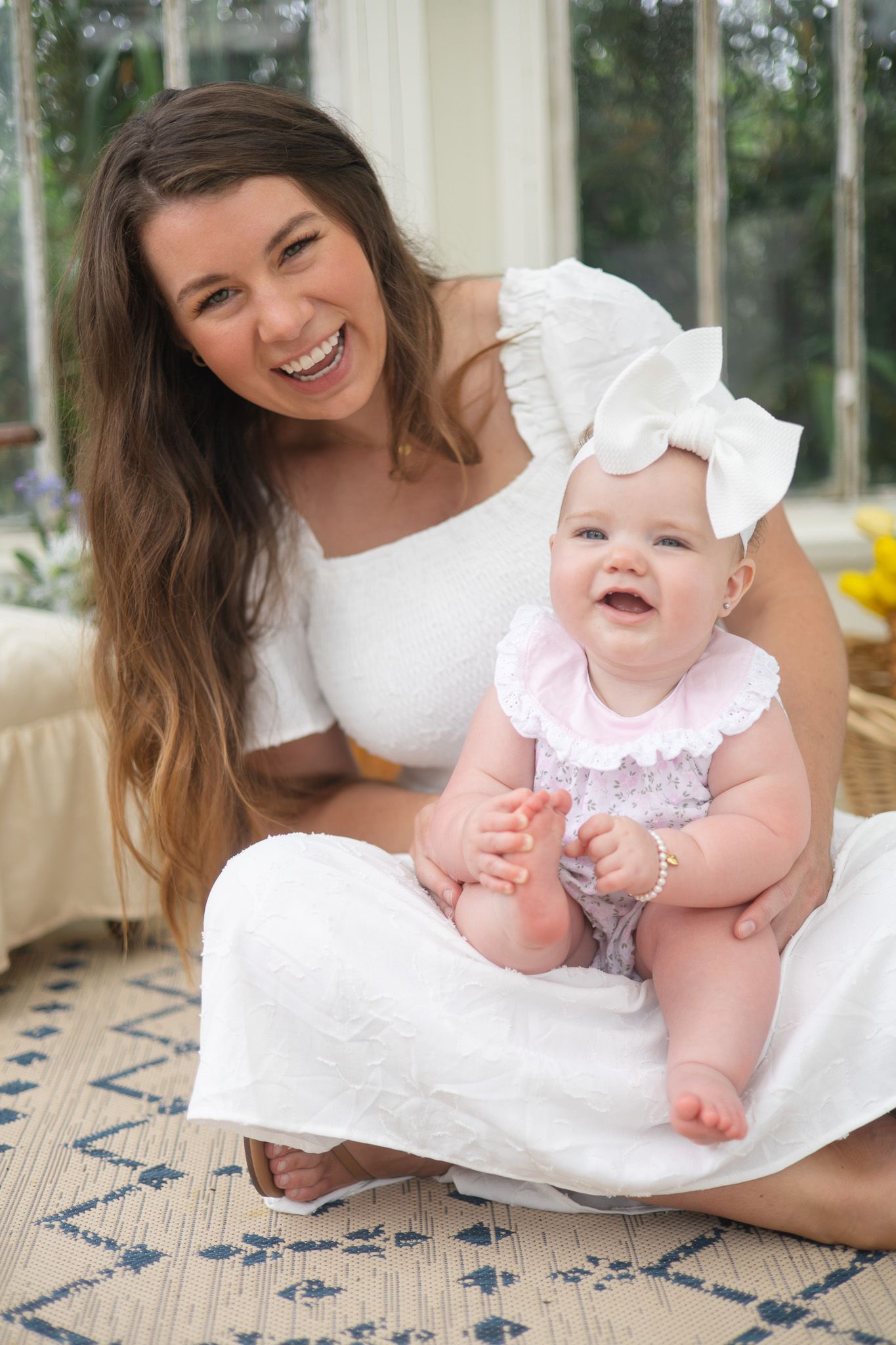 Laughing young mother in white dress holds smiling baby girl in pink romper with bow.