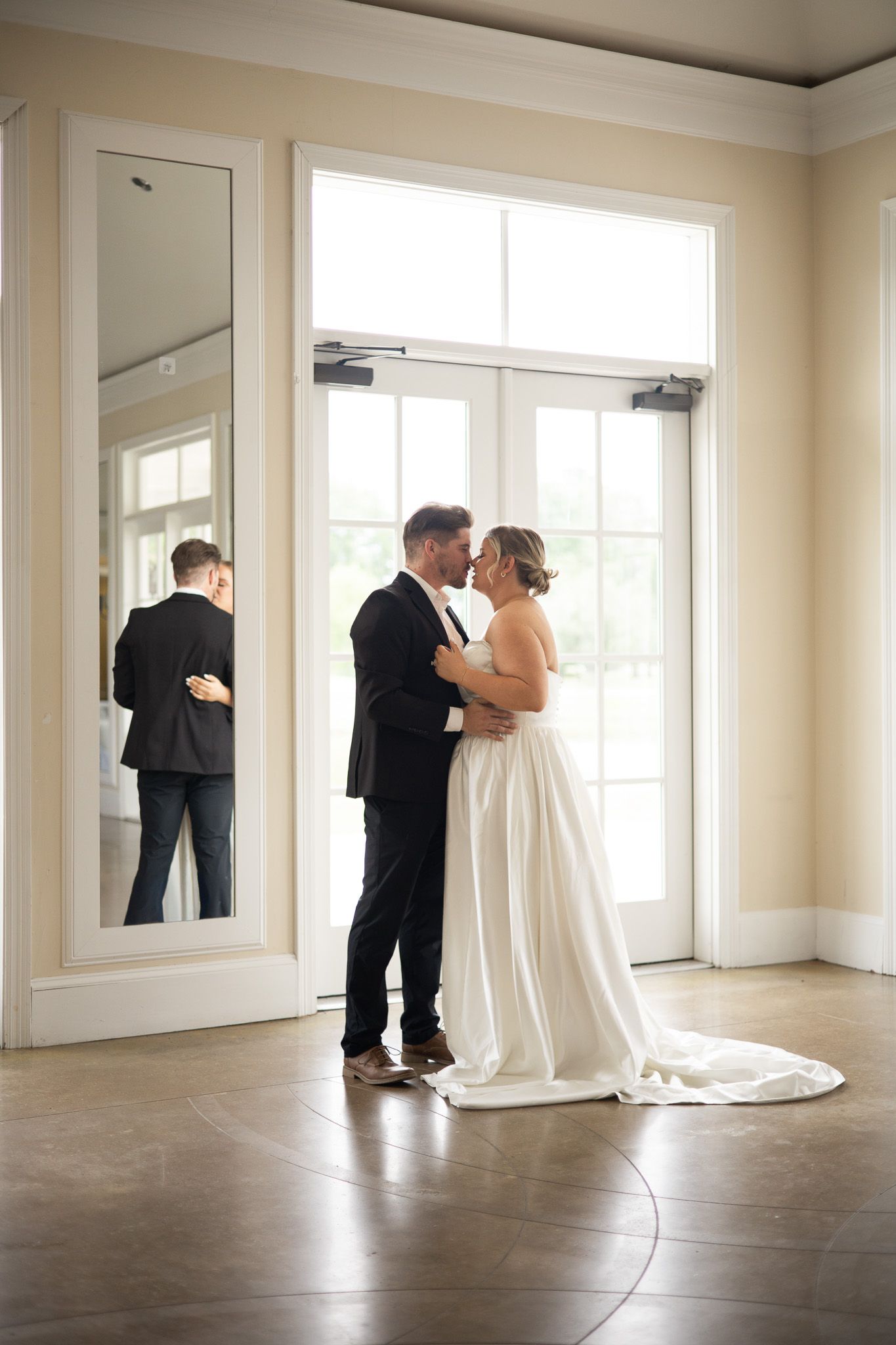 Bride and groom sharing a romantic embrace near French doors, their reflection visible in a large mirror nearby.