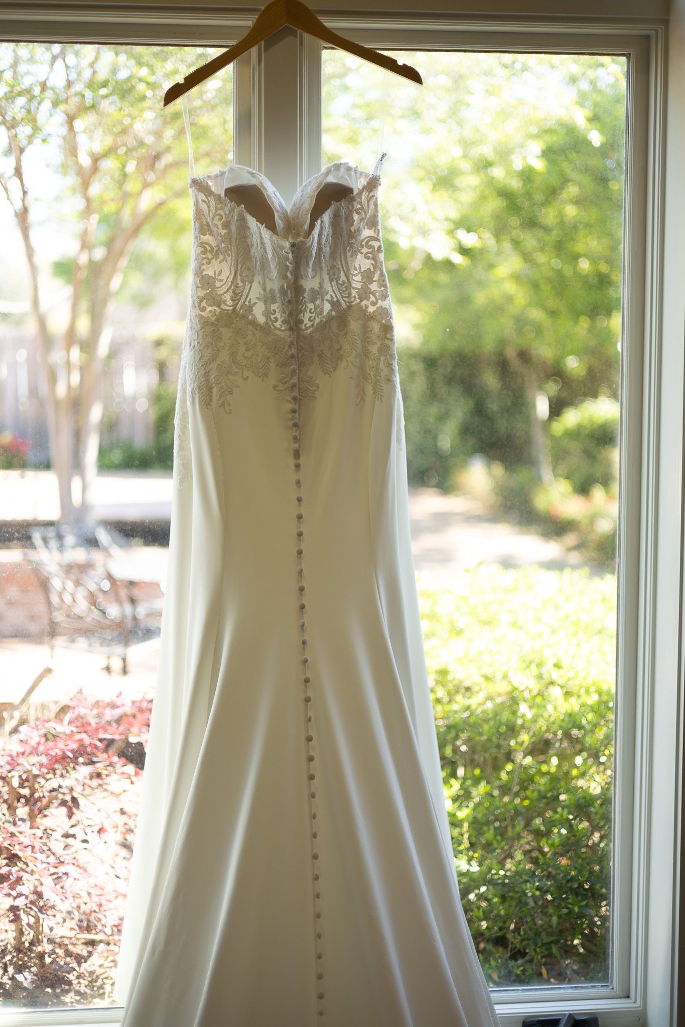 Elegant white strapless mermaid wedding dress with lace bodice and button back, hanging in a sunlit window.