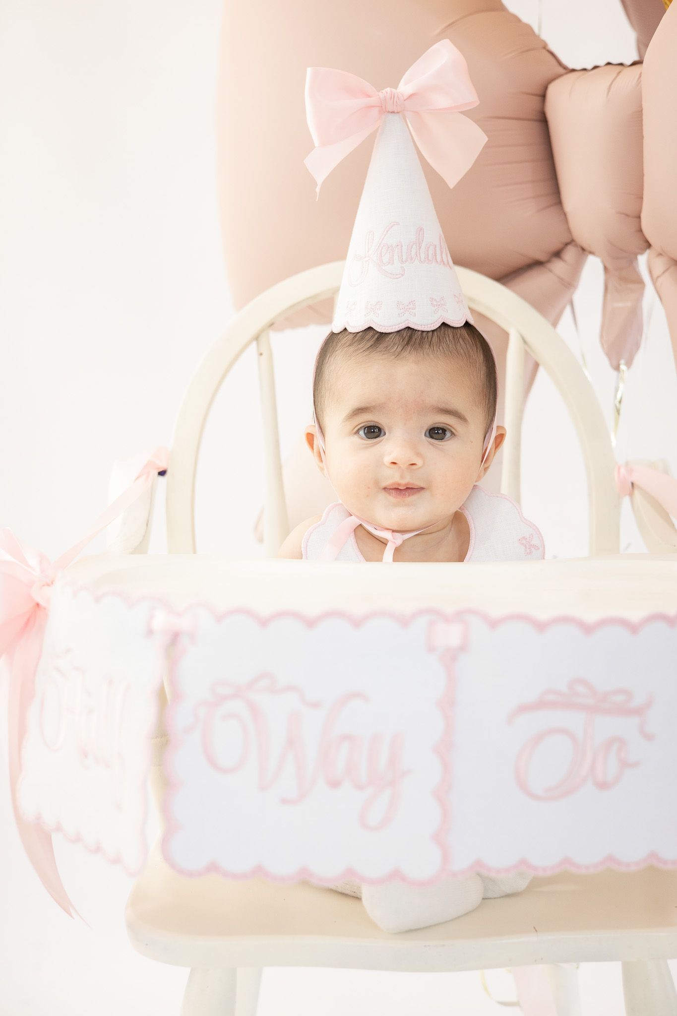 Adorable baby girl in a pink birthday hat sits in a high chair decorated with a pink banner for her first birthday.