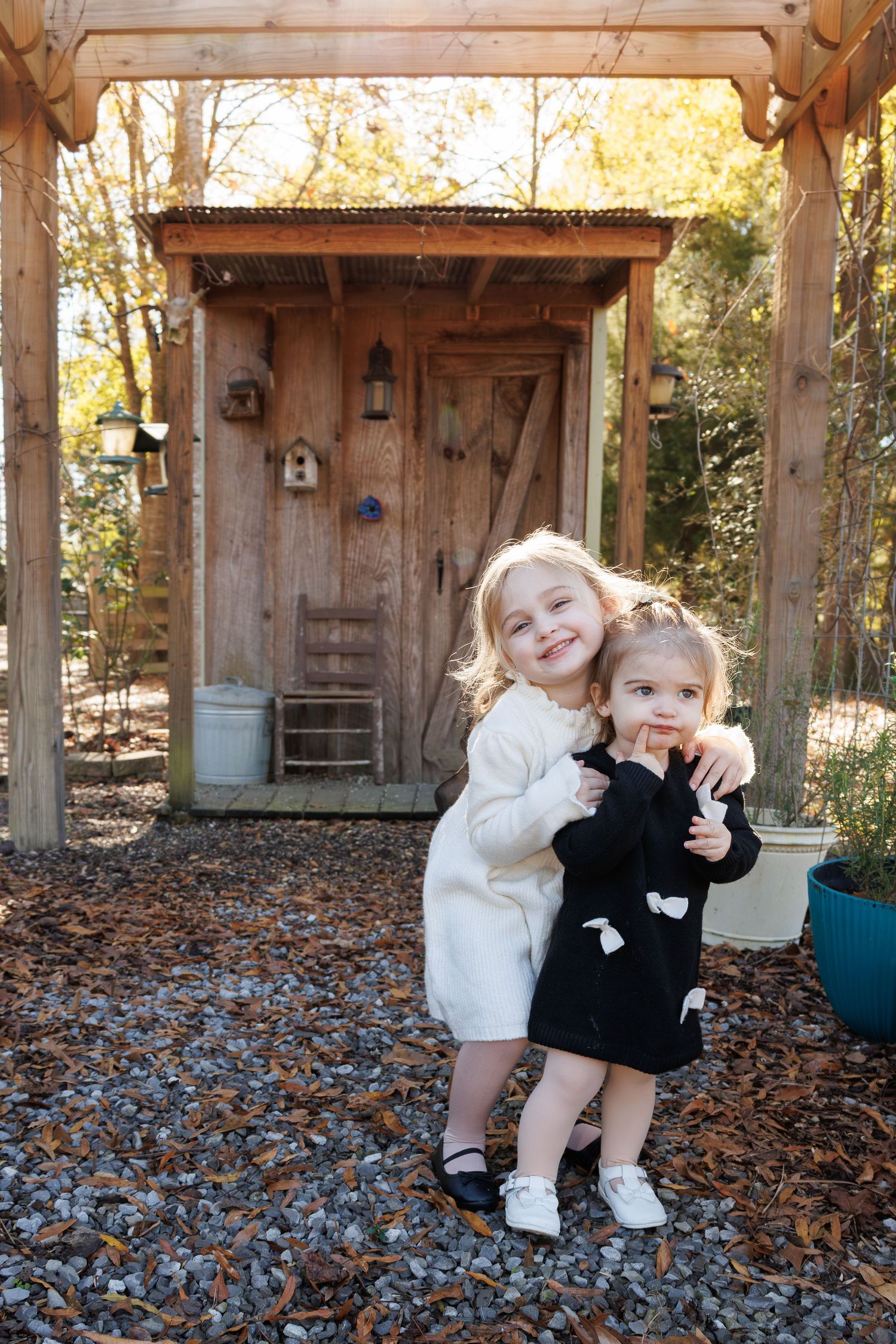 Two young girls hugging and smiling outdoors in front of a rustic wooden shed.