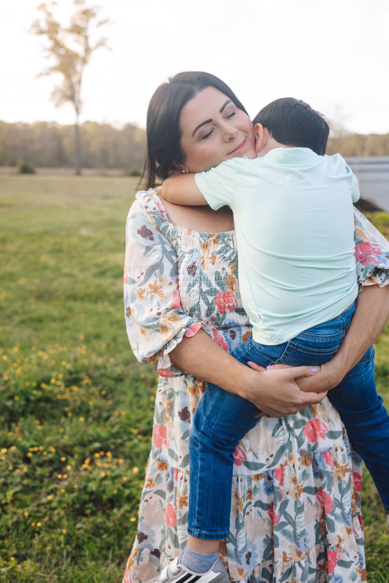 Mother in floral dress hugging young child outdoors in green field at golden hour.