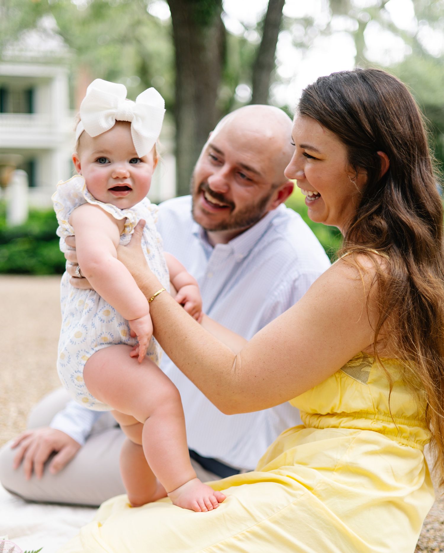 A happy family with a bald father, brunette mother in yellow dress, and baby girl with white bow sitting outdoors.