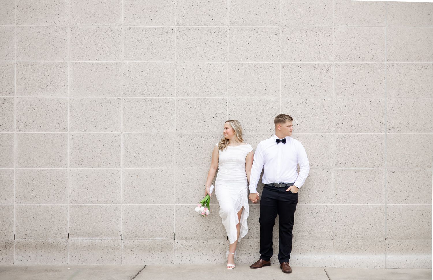 Bride in white dress holding bouquet and groom in black tuxedo hold hands against a stone wall.