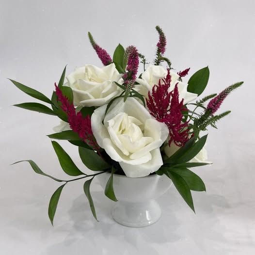 White roses and burgundy veronica blooms arranged in a white ceramic vase with lush green foliage.