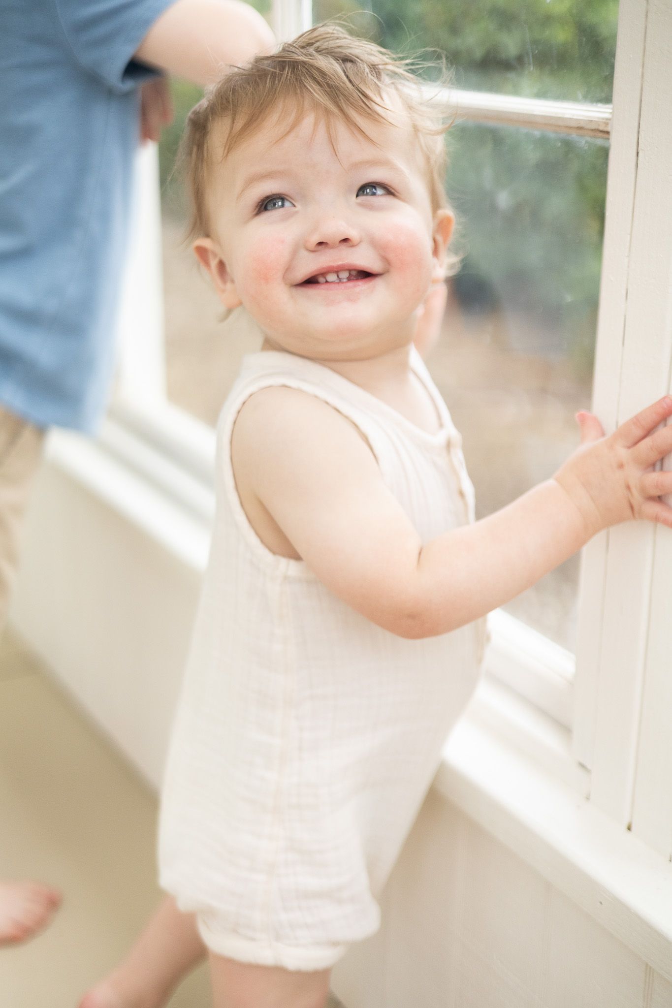 A happy toddler in a white romper stands by a bright window, smiling and holding onto the frame.