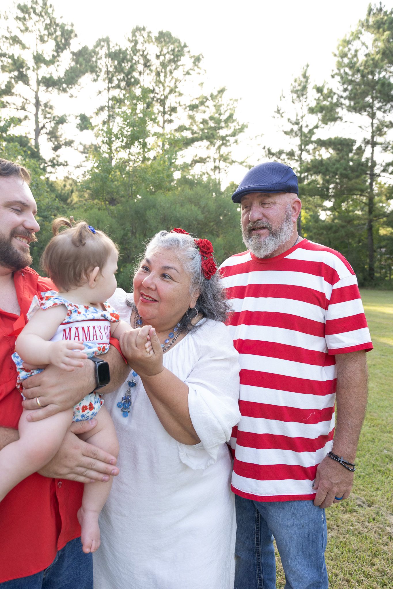 Multi-generational family outdoors, grandparents with adult son holding baby, wearing red and white outfits in a green park setting.