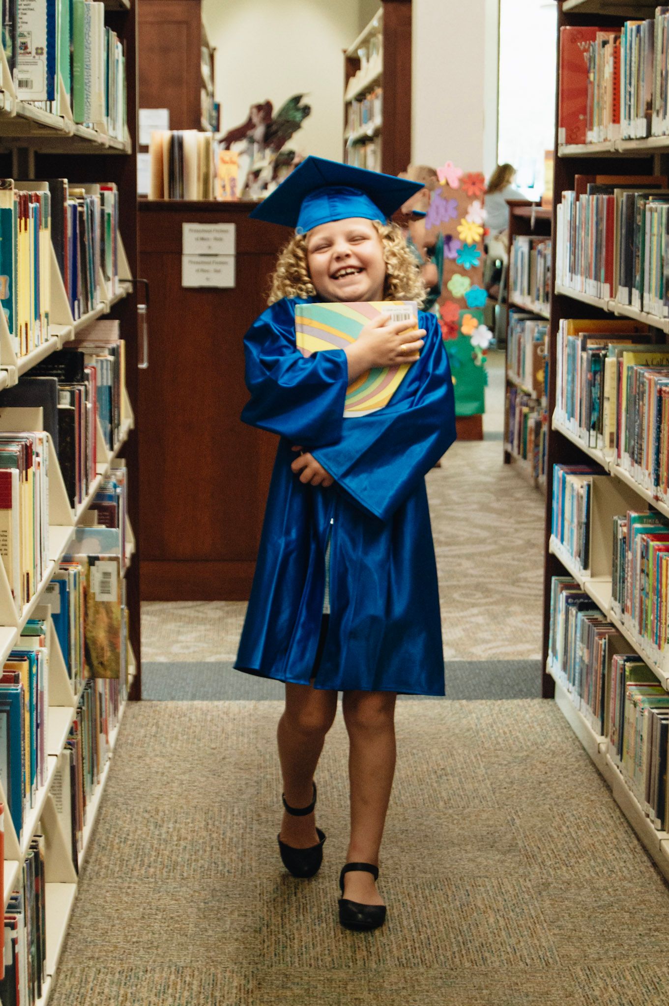 Happy young girl in blue graduation cap and gown holding a book in a library aisle.