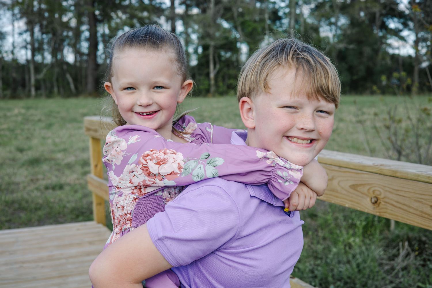 Two young siblings smile and hug on a wooden deck with green trees in the background.